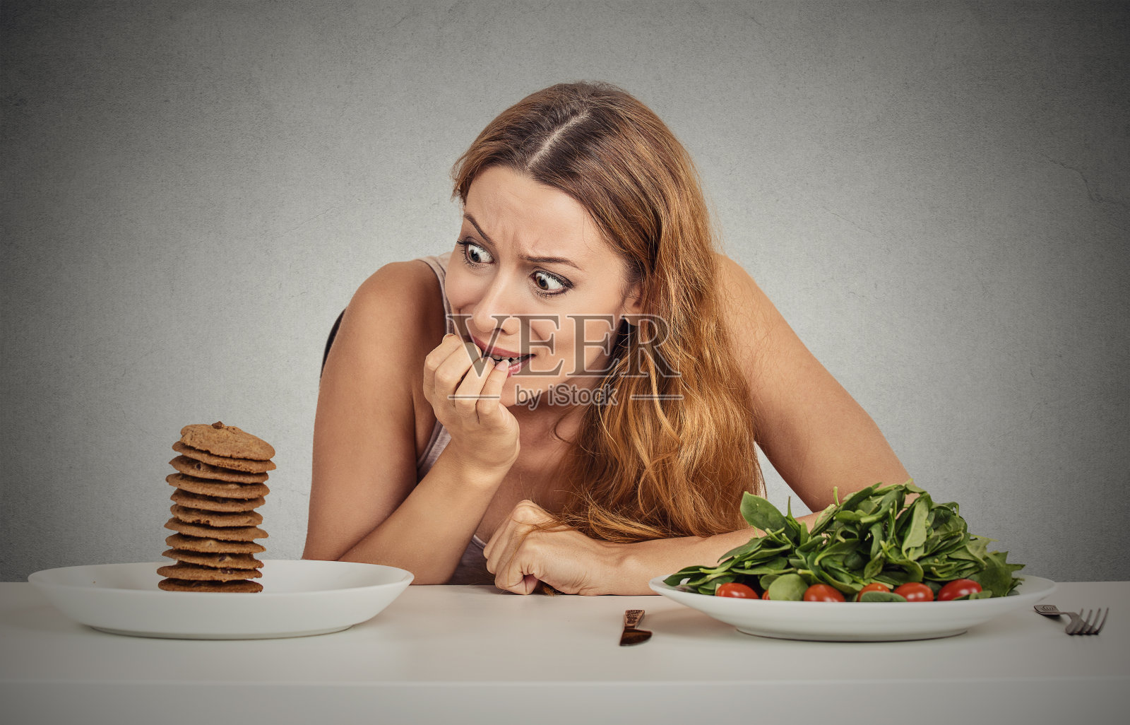 年轻女子坐在餐桌前决定是吃健康食品还是她想吃的甜饼干照片摄影图片