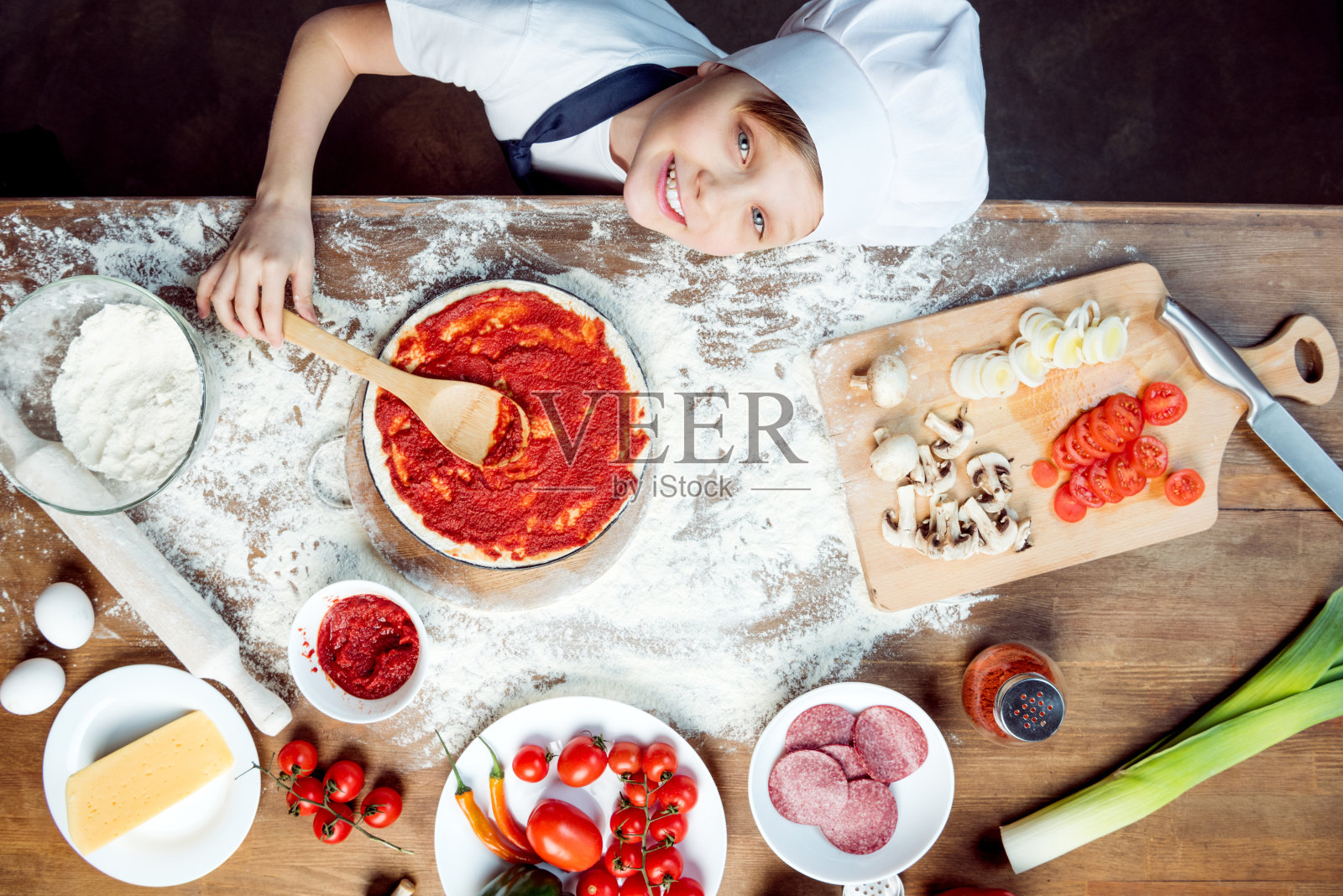 俯视图的男孩做比萨饼的材料，比萨饼，西红柿，意大利腊肠和蘑菇在木制的桌面上照片摄影图片