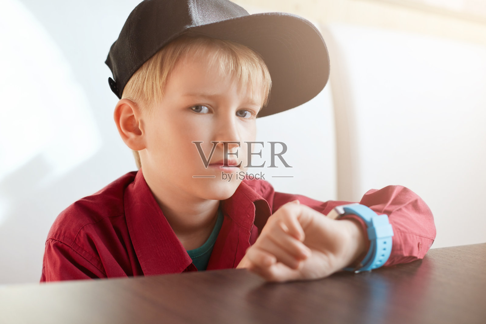 一个严肃的男孩戴着时髦的帽子，穿着红色的衬衫，手腕上戴着智能手表，坐在白色背景的木桌前的横向肖像。一个英俊的小男孩带着手表。照片摄影图片