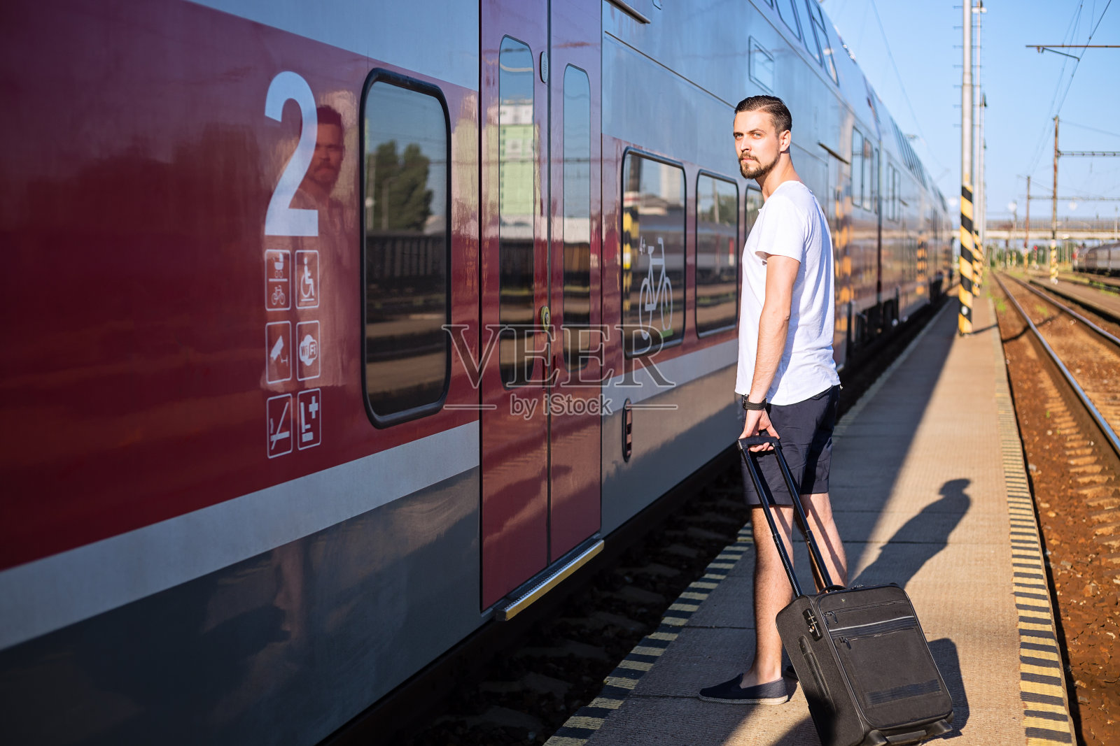 一个白种男人带着手提箱站在火车旁边，穿着休闲的夏装。照片摄影图片