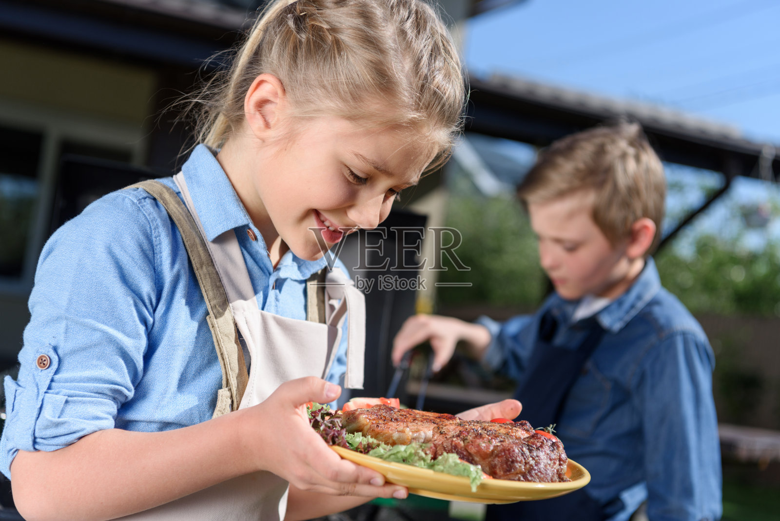 可爱的小女孩拿着一盘新鲜的牛排，而她的弟弟正在准备烤肉照片摄影图片