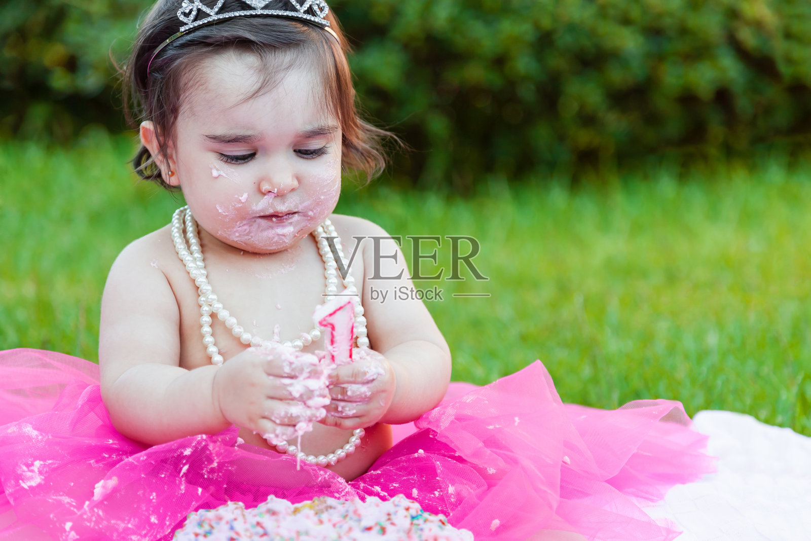 蹒跚学步的小女孩在一周年生日派对上玩蜡烛，弄脏了脸，用蜡烛做了一团粉红色的蛋糕。照片摄影图片