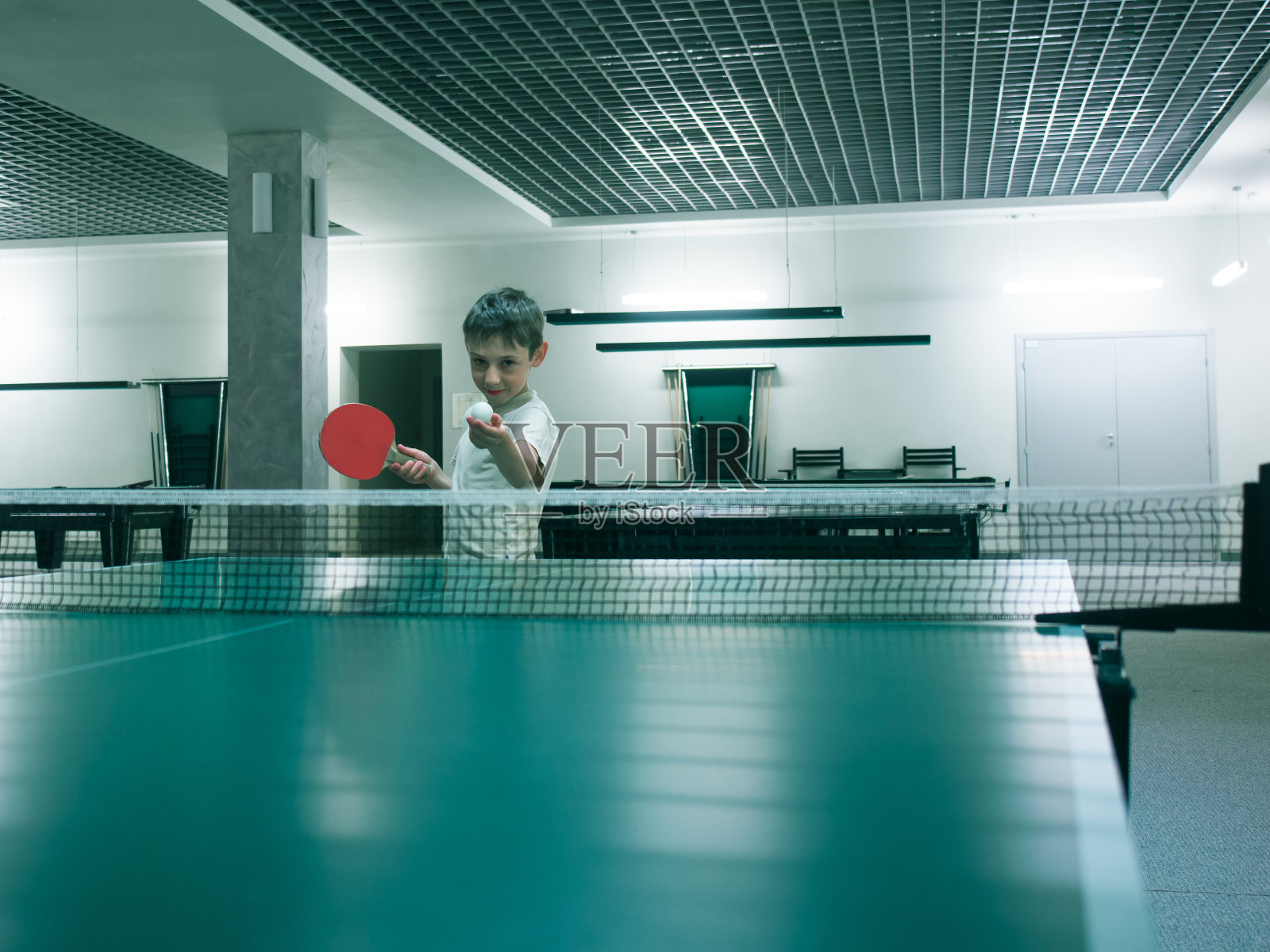 一个小男孩在一件t恤谁拿着网球球拍和球在网球桌上和空房间色调的形象照片摄影图片