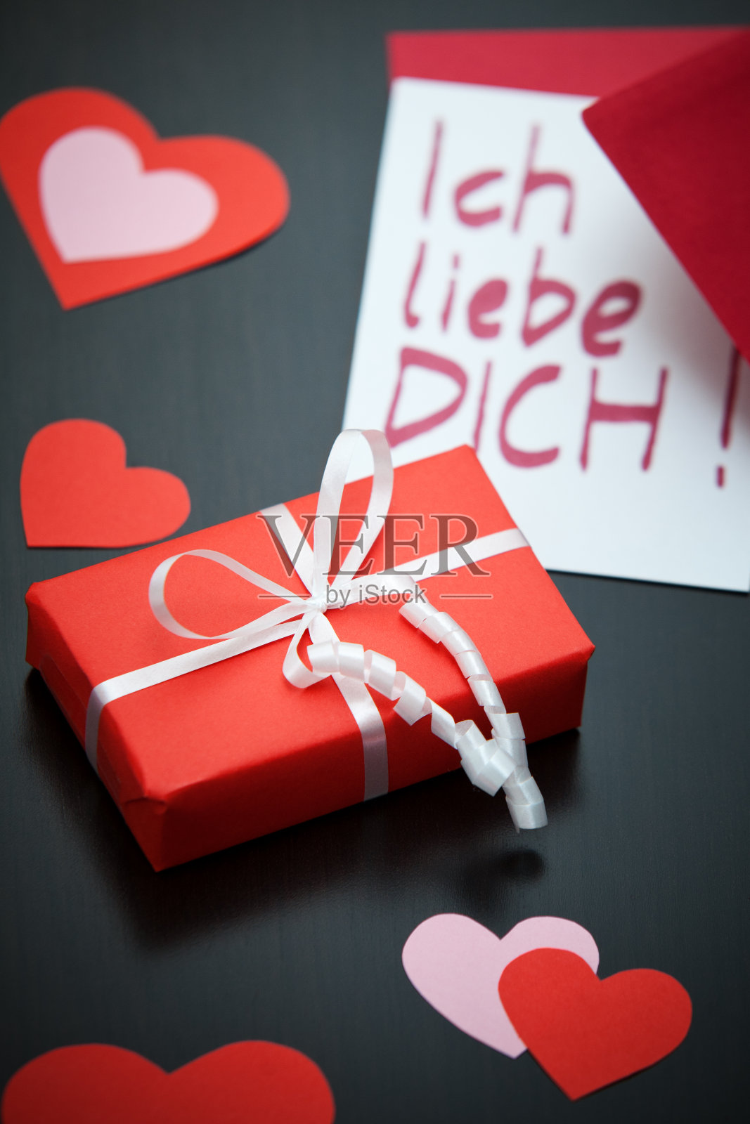 黑色的桌子上放着一个带白色丝带的红色包裹和一张带有红色信封的“我爱你”(德语)纸条，包裹周围是红粉色的纸心。照片摄影图片