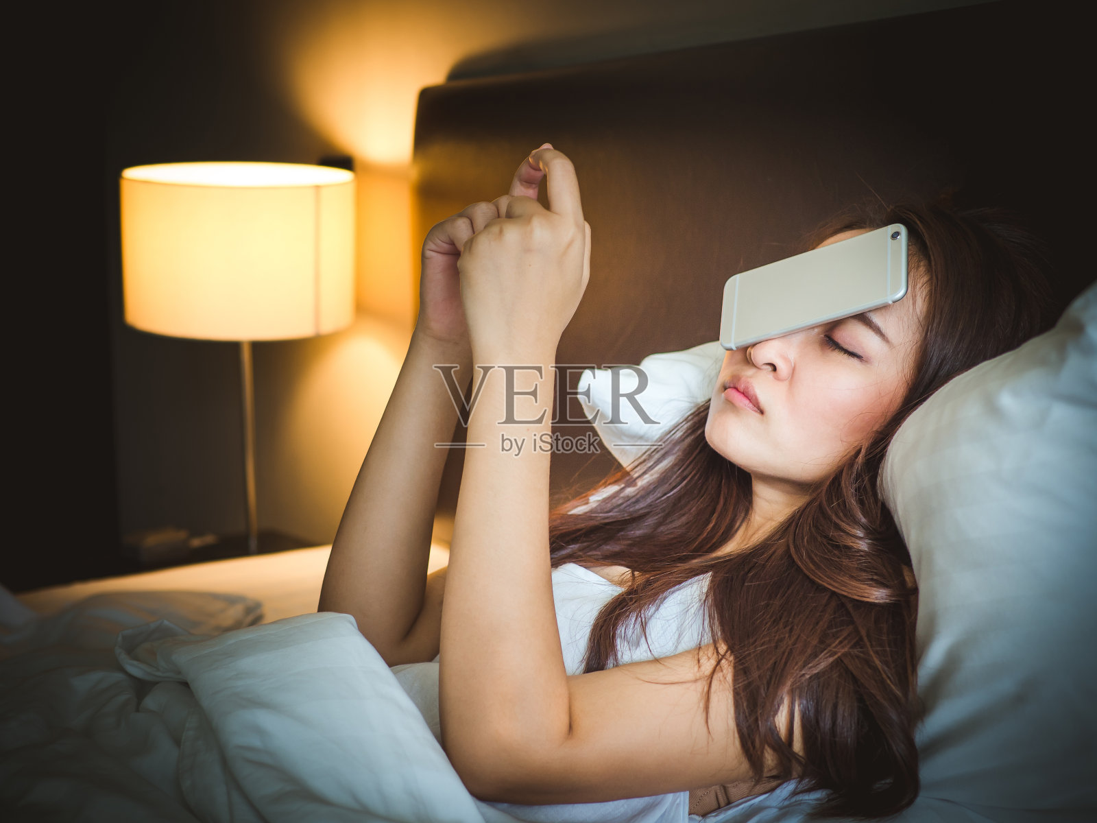 睡在白色床上的亚洲女人，把智能手机掉在脸上照片摄影图片