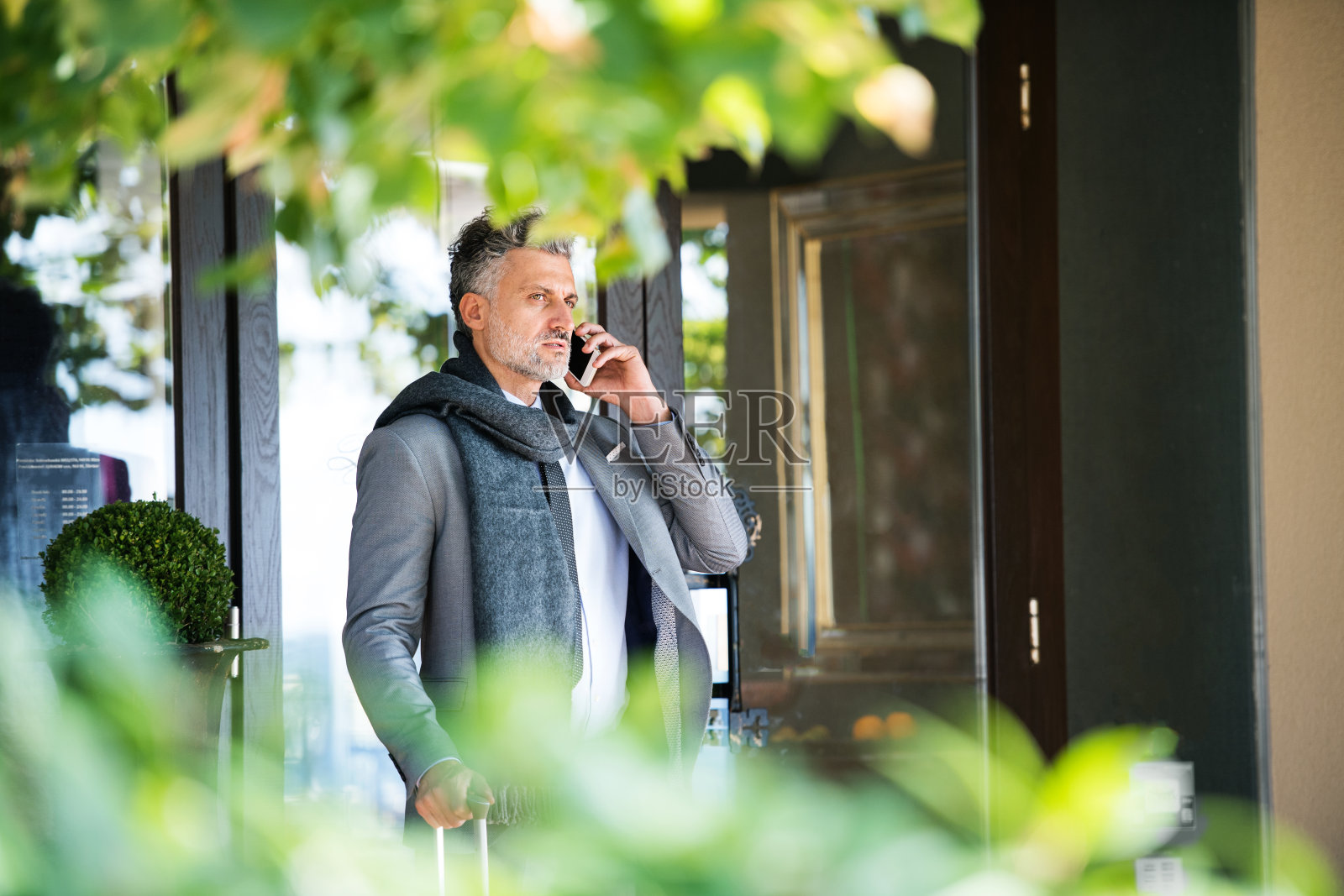 一个拿着智能手机的成熟商人站在酒店前。照片摄影图片
