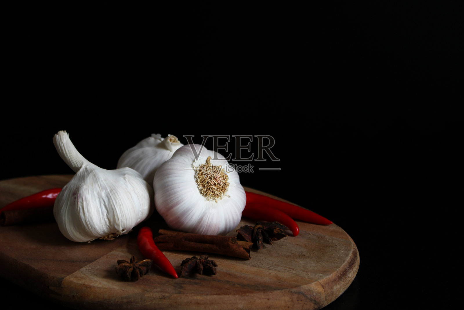 厨房的静物画中有生大蒜、红辣椒、桂皮和八角香料放在木砧板上，黑色背景。大蒜的蔬菜照片摄影图片
