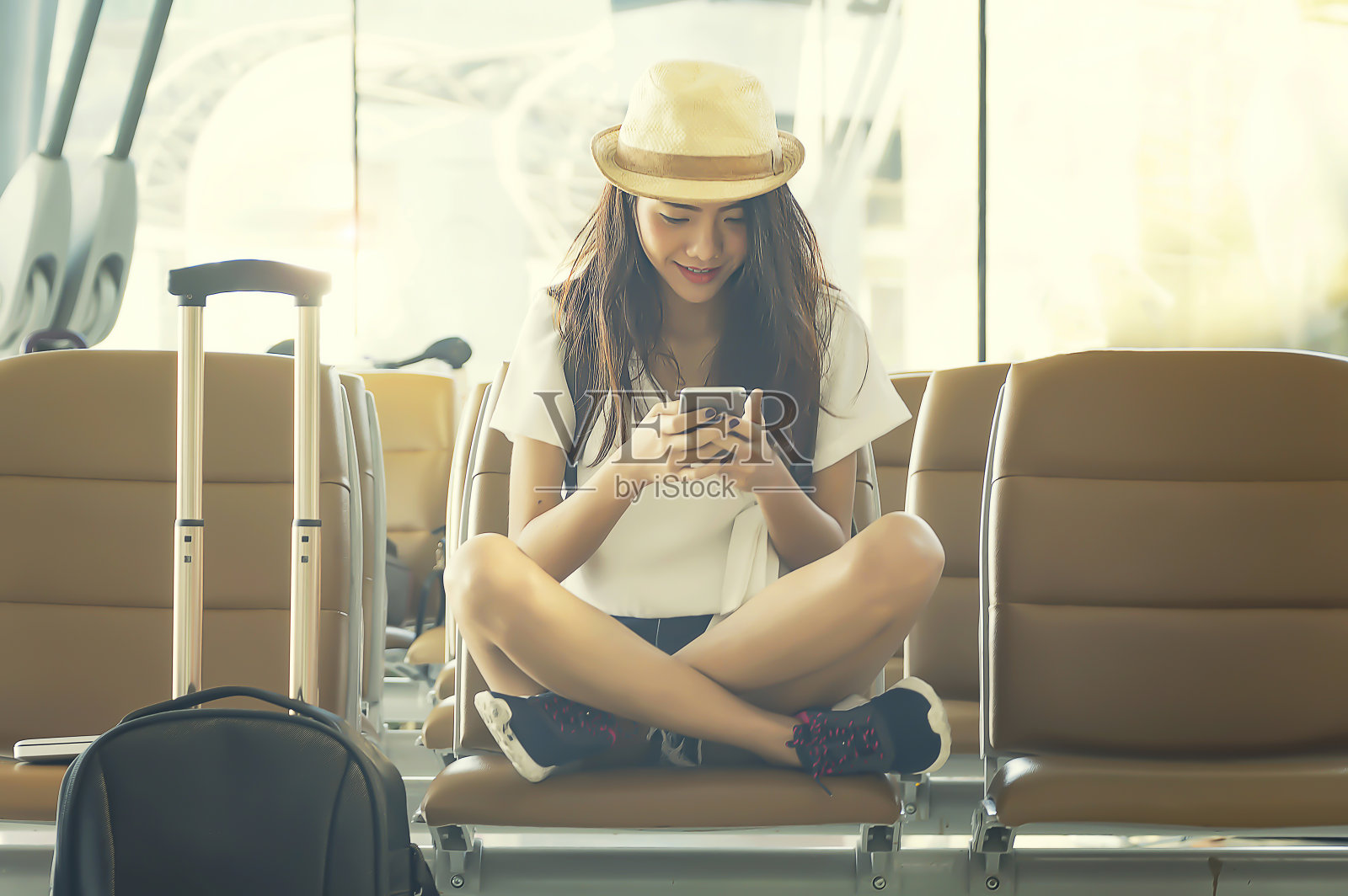 年轻女子在机场用智能手机等待飞机。她推着旅行箱手推车坐在机场候机室的候机室里。旅行的概念。照片摄影图片