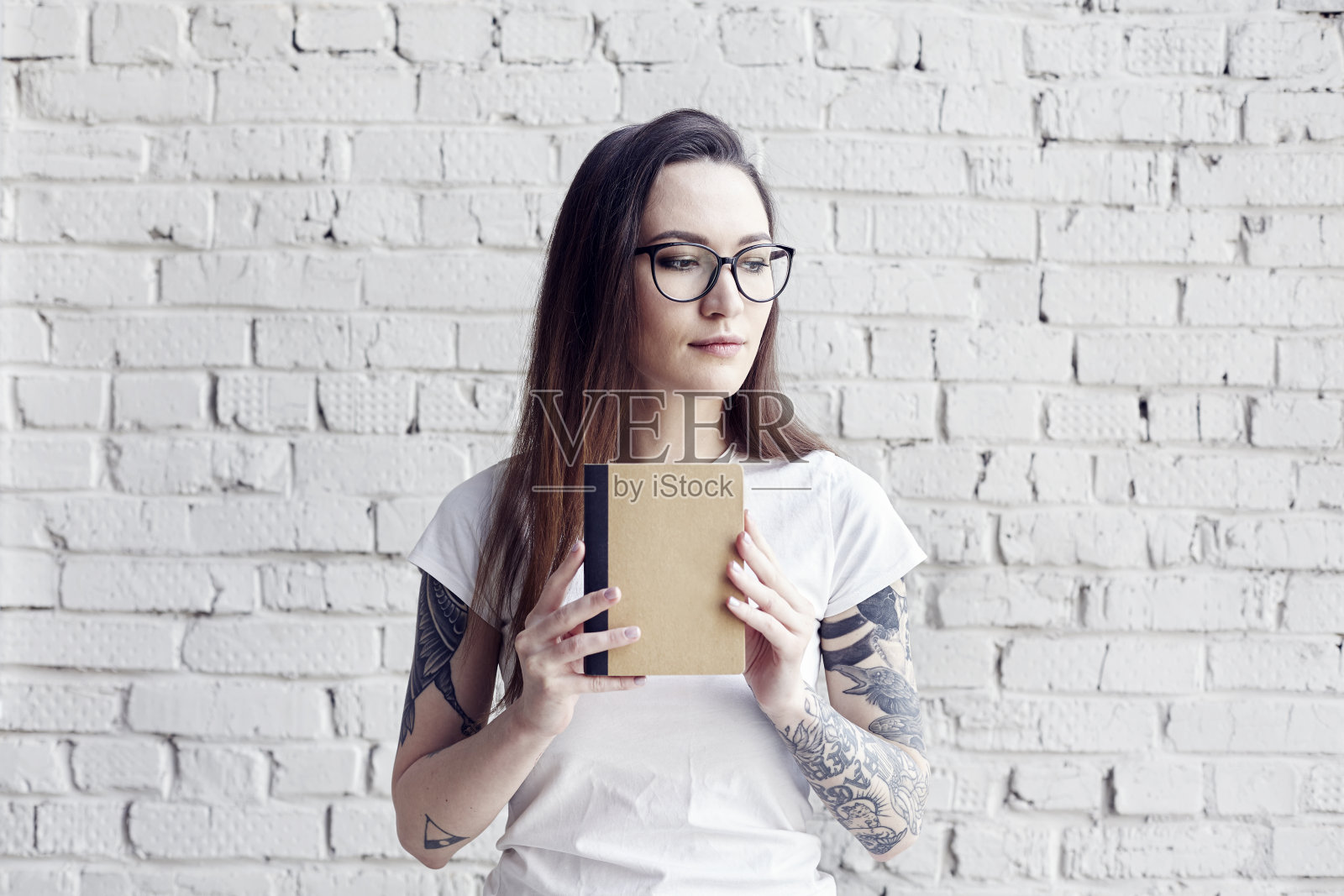 漂亮的潮人纹身女子摆姿势白色t恤与书在手里，孤立在白色砖墙，手持一本书，设计布局的空间照片摄影图片