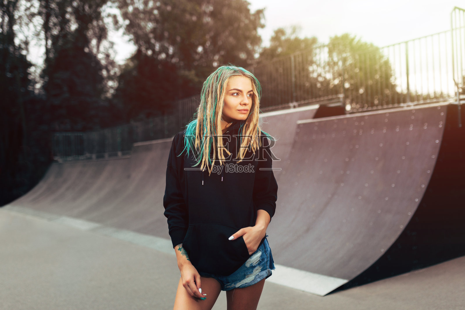 美丽年轻的健身女人在黑色帽衫站在滑板公园。照片摄影图片