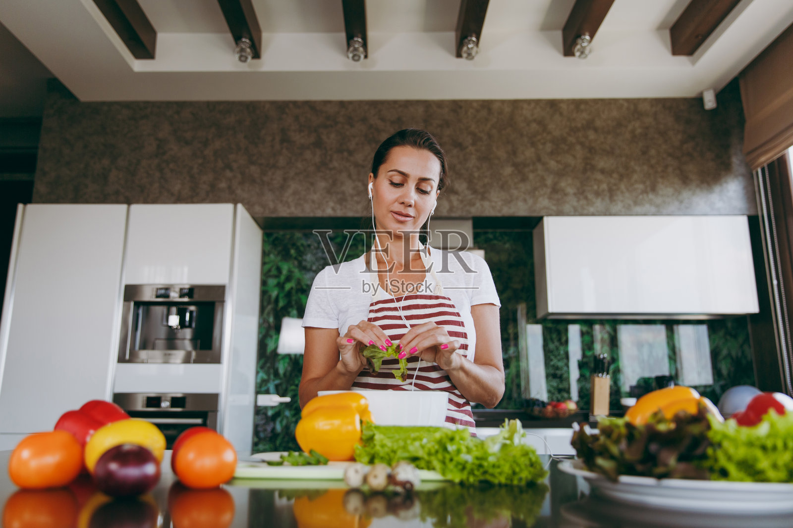 年轻女子戴着耳机，手里拿着蔬菜，在厨房桌子上放着笔记本电脑。蔬菜沙拉。节食的概念。健康的生活方式。在家做饭。准备食物照片摄影图片