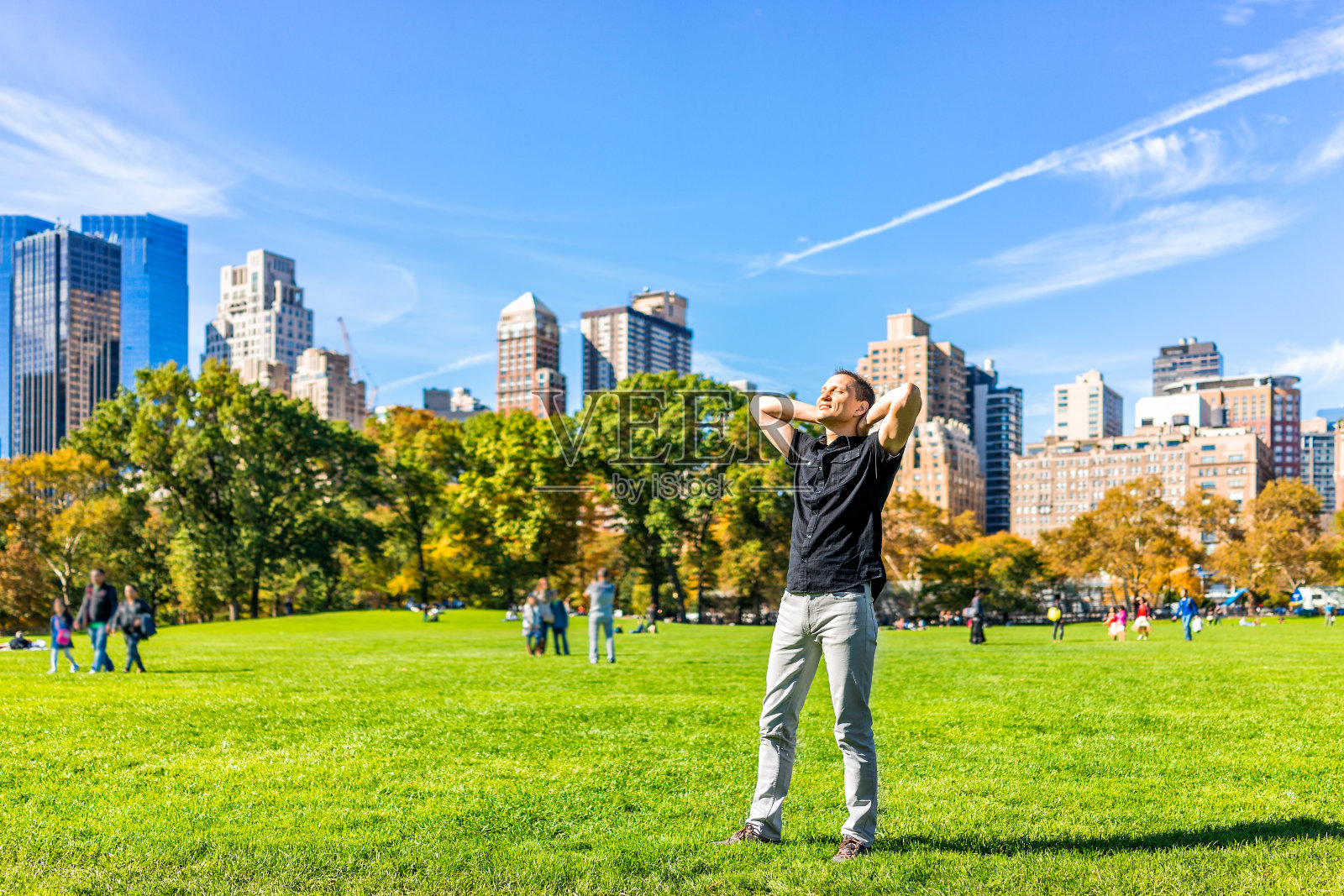 一个快乐的年轻人举起手在纽约中央公园在阳光明媚的秋日与现代摩天大楼和人的特写照片摄影图片