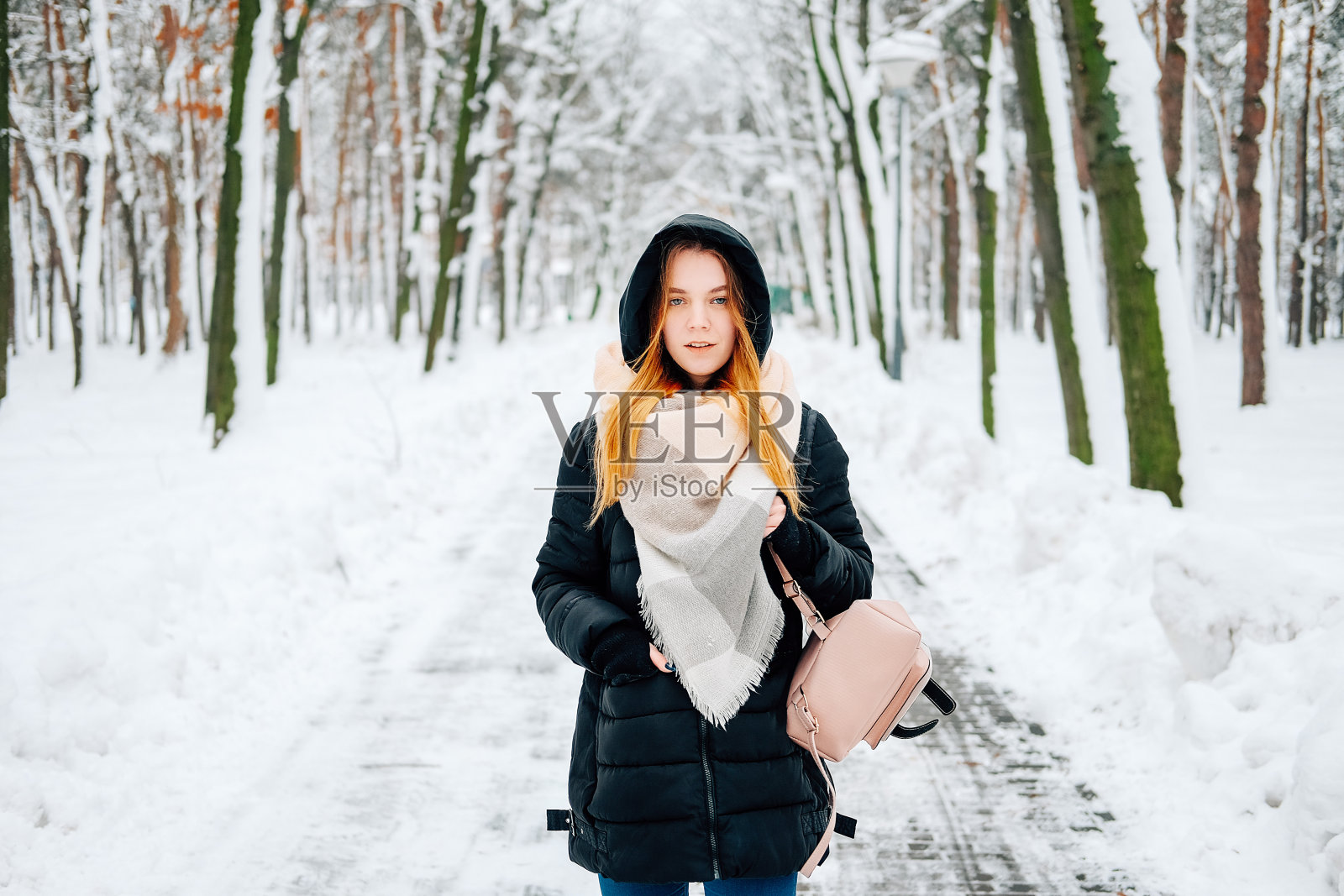 迷人的金发年轻女子走在冬天的森林里满是雪，穿着黑色皮大衣，牛仔裤，粉色皮靴，背包和米色披肩的休闲服装照片摄影图片