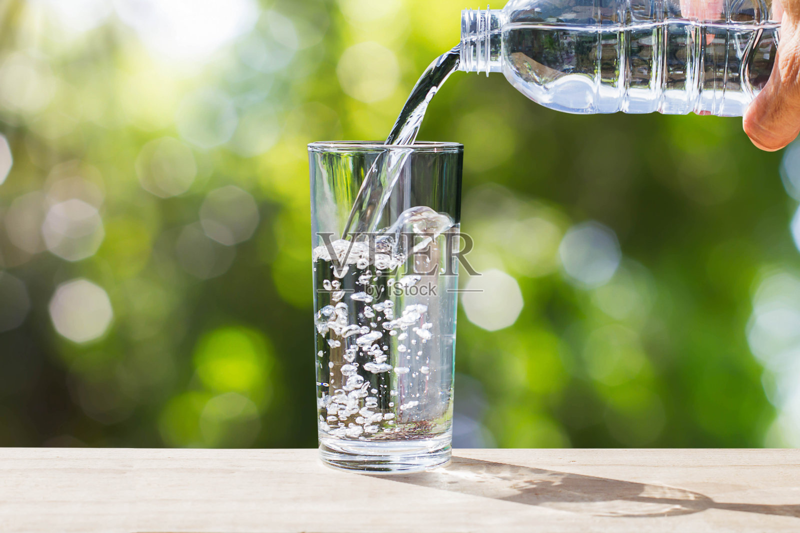 手握水瓶，将水倒进木质桌面的玻璃中，背景是模糊的绿色散景，阳光柔和照片摄影图片