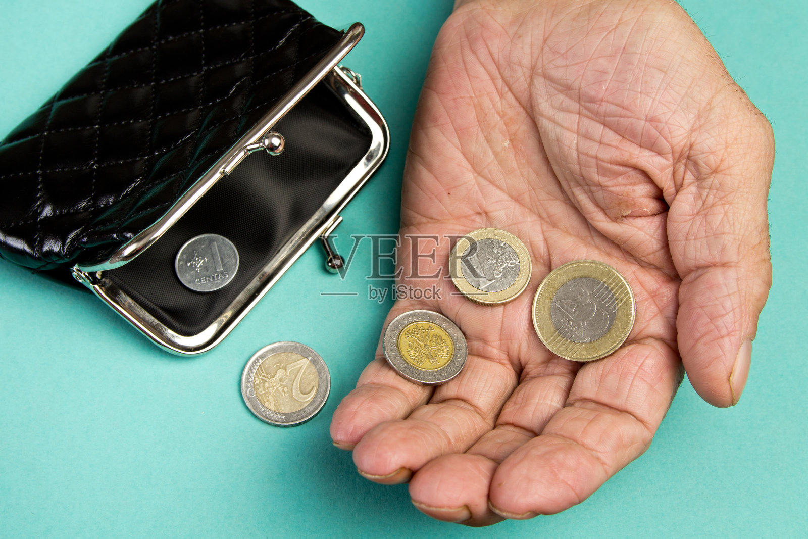 一个老人把硬币放在空空的旧皮夹上。退休后贫困的概念。黑色钱包的变化在绿松石背景照片摄影图片