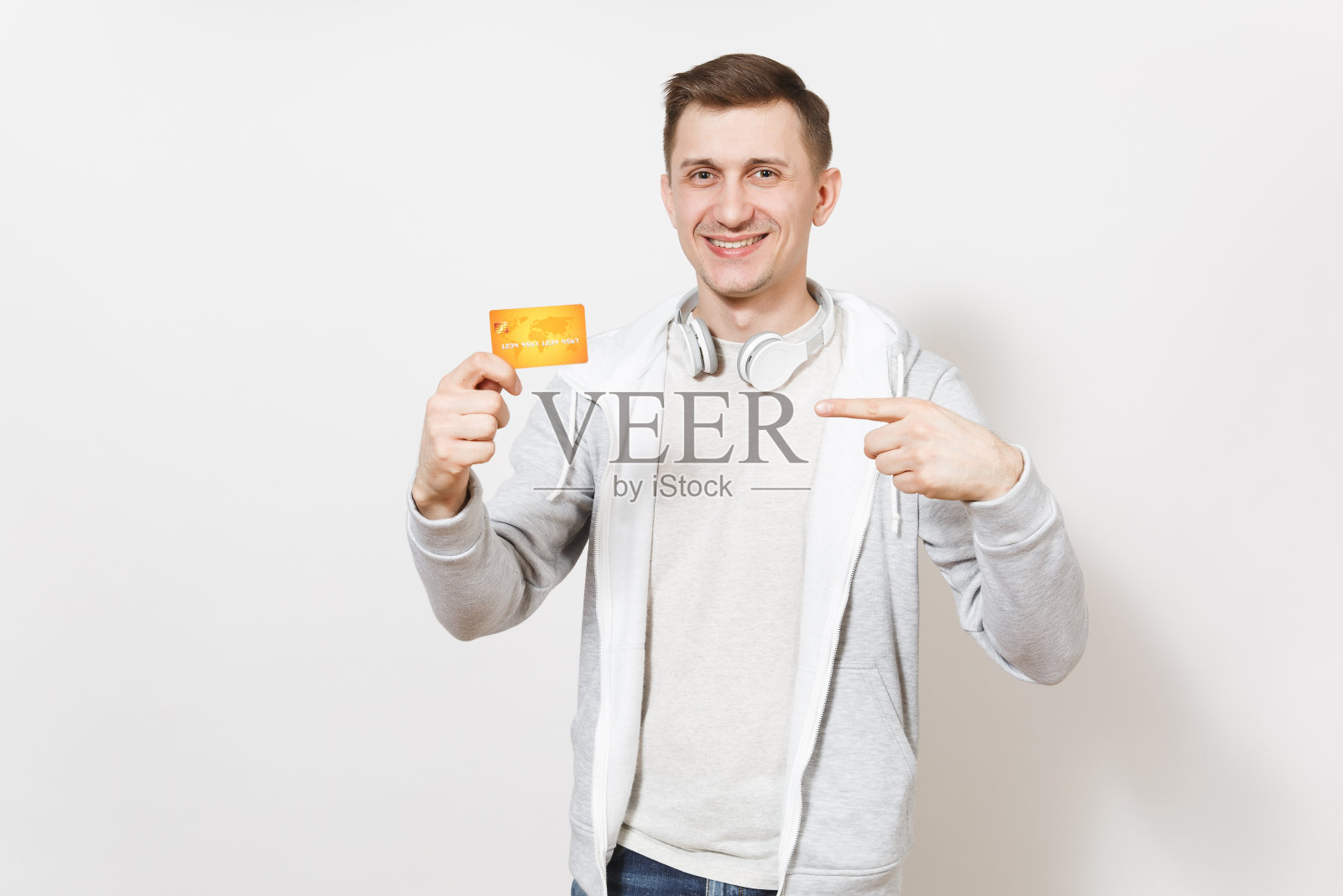 年轻英俊的微笑学生在t恤和轻运动衫脖子上戴着耳机拿着橙色的信用卡，用手指指着它在工作室的白色背景。成功的概念照片摄影图片