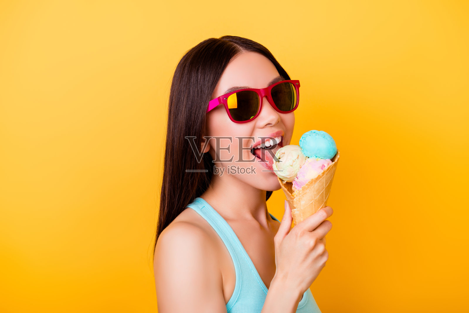 开朗的年轻韩国小姐舔着三勺不同口味的美味冰淇淋，站在黄色的背景上，戴着旅游眼镜照片摄影图片