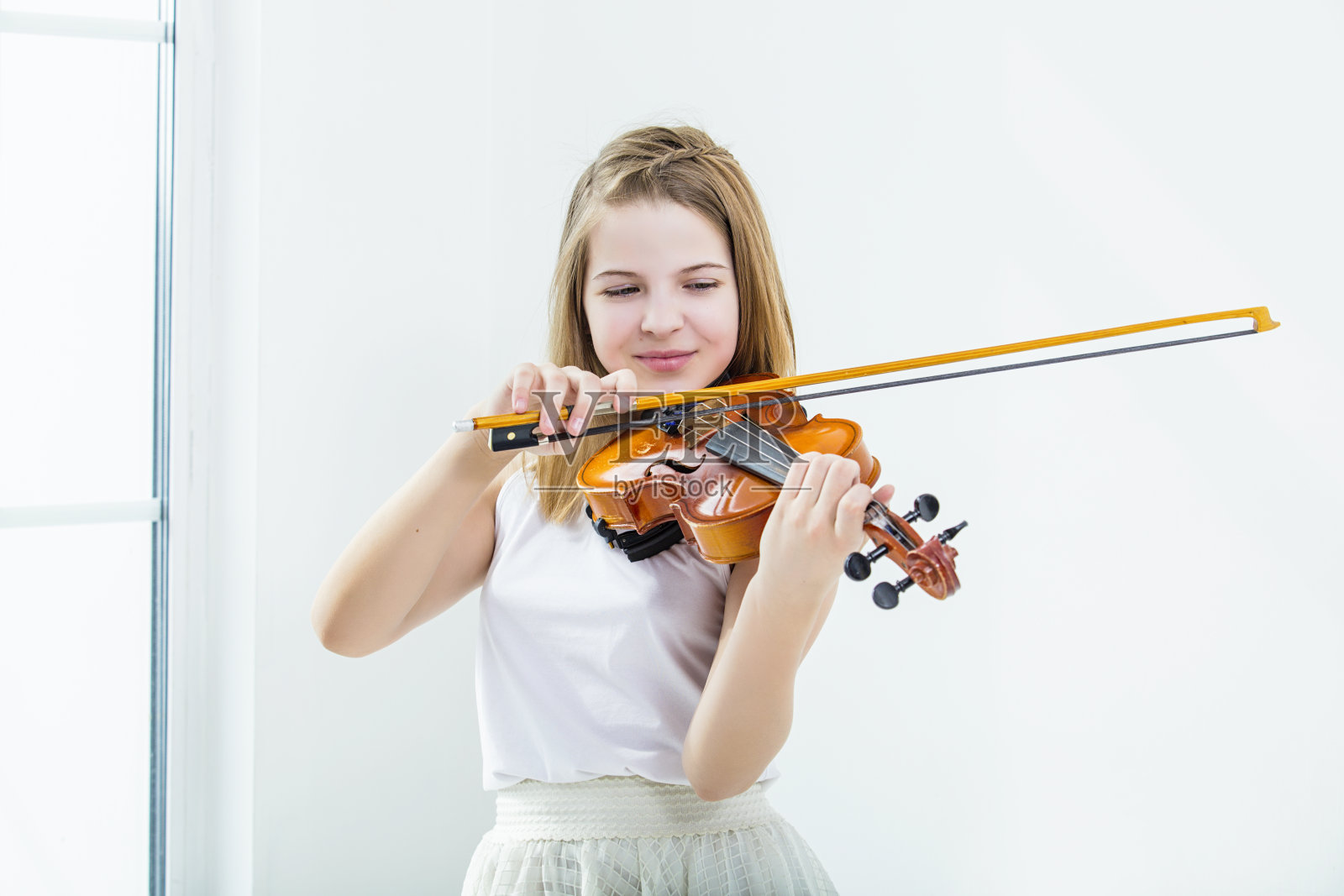 小女孩在一间有窗户的白色房间里拉着小提琴学习，美丽而快乐照片摄影图片