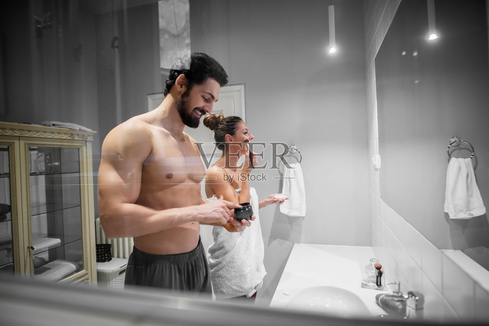 年轻美丽迷人的女孩与她赤裸的肌肉英俊的男朋友或丈夫站在浴室毛巾和准备他们的皮肤和头发一起在镜子前。照片摄影图片