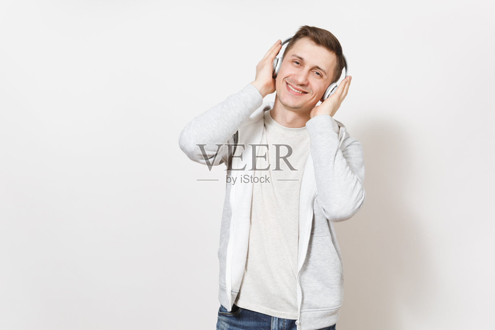 年轻英俊的微笑快乐的男人学生在t恤和轻运动衫听音乐与白色无线耳机，把他们的手孤立在白色背景。情感的概念照片摄影图片