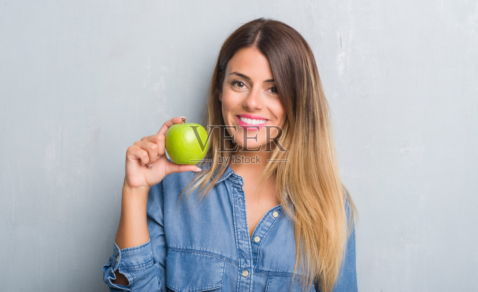 年轻的成年女子越过灰色的垃圾墙吃新鲜的绿苹果，快乐的脸站着，微笑着自信的微笑显示牙齿照片摄影图片