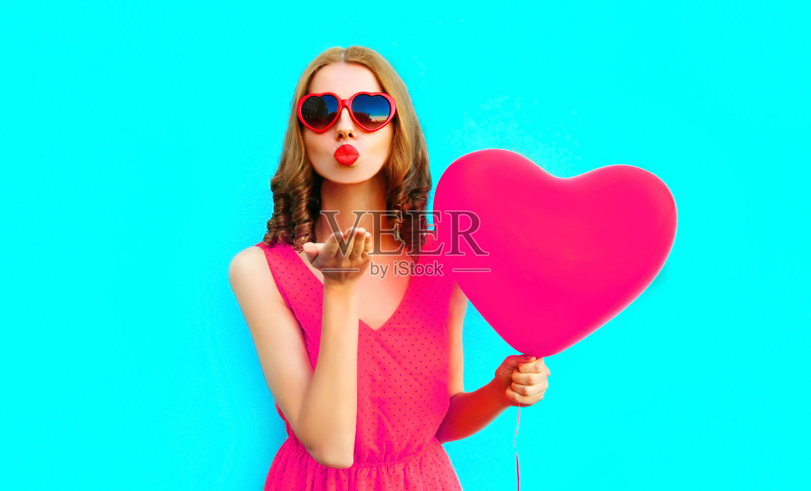 在彩色的蓝色背景上，年轻女子用粉红色的心型气球送出甜蜜的飞吻照片摄影图片