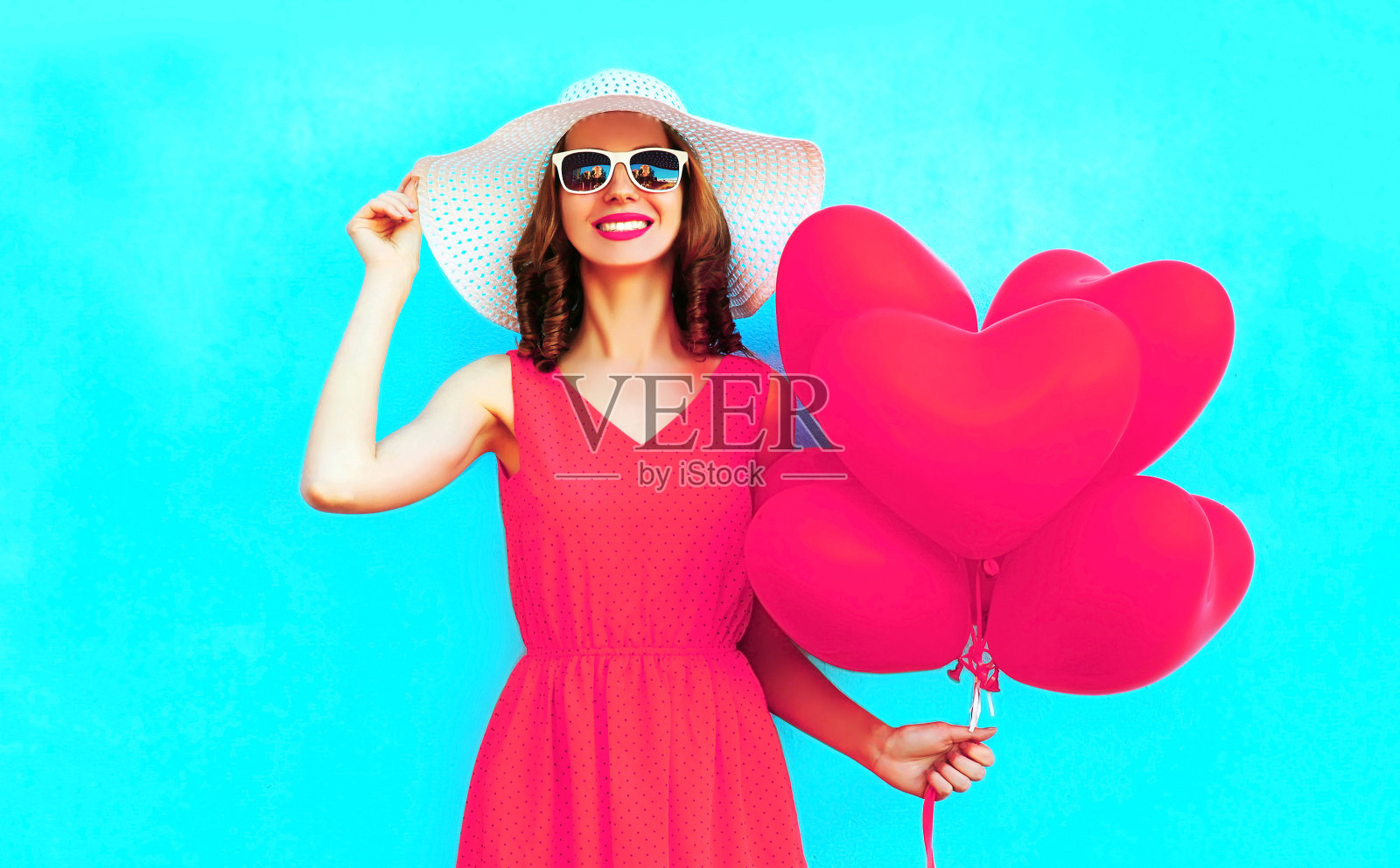 肖像快乐微笑的妇女手持粉红色的心形气球在蓝色的背景，戴着夏季草帽照片摄影图片