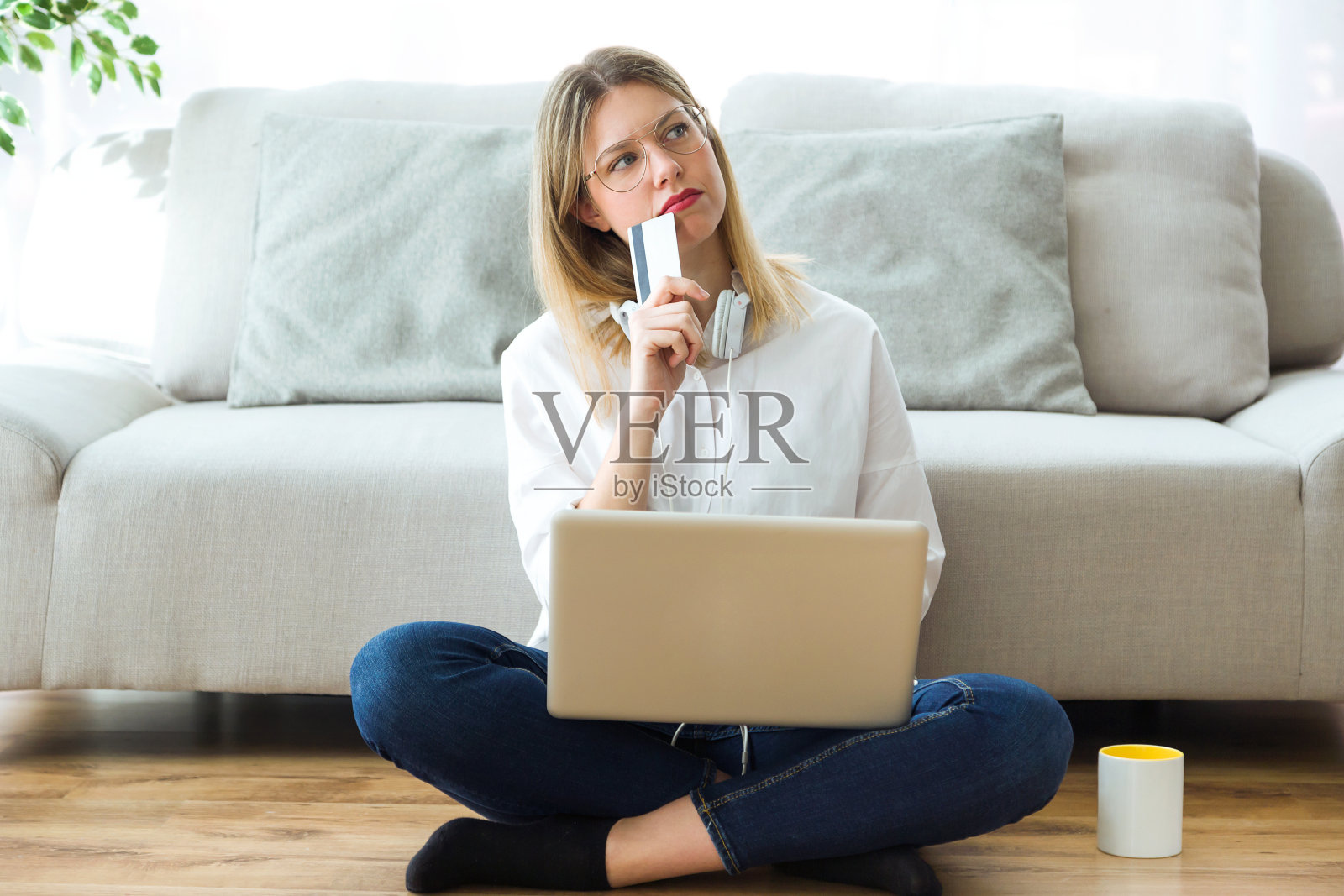 年轻漂亮的女人坐在家里的地板上想着网上购物或不购物。照片摄影图片