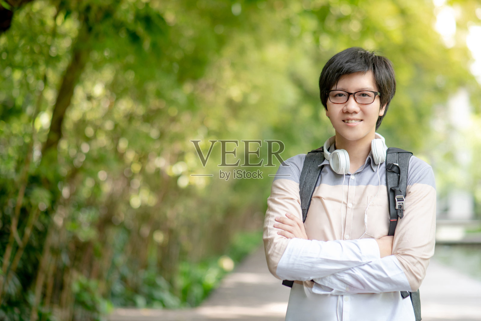 自信的亚洲人大学学生戴着眼镜和耳机微笑和交叉手臂在绿色的大学公园。快乐的校园生活。教育机会和奖学金的概念照片摄影图片
