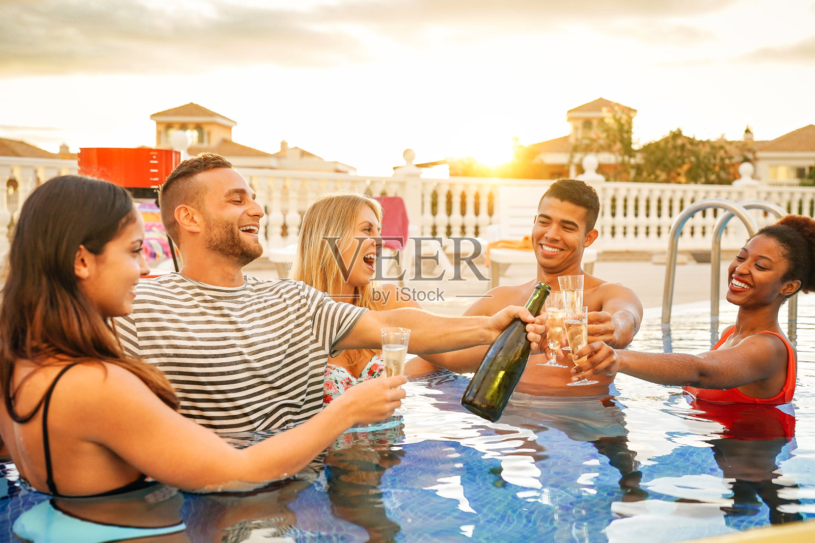 一群快乐的朋友使一个游泳池派对与一瓶香槟在日落-年轻人有乐趣喝起泡酒在豪华的热带度假-青年的生活方式概念照片摄影图片