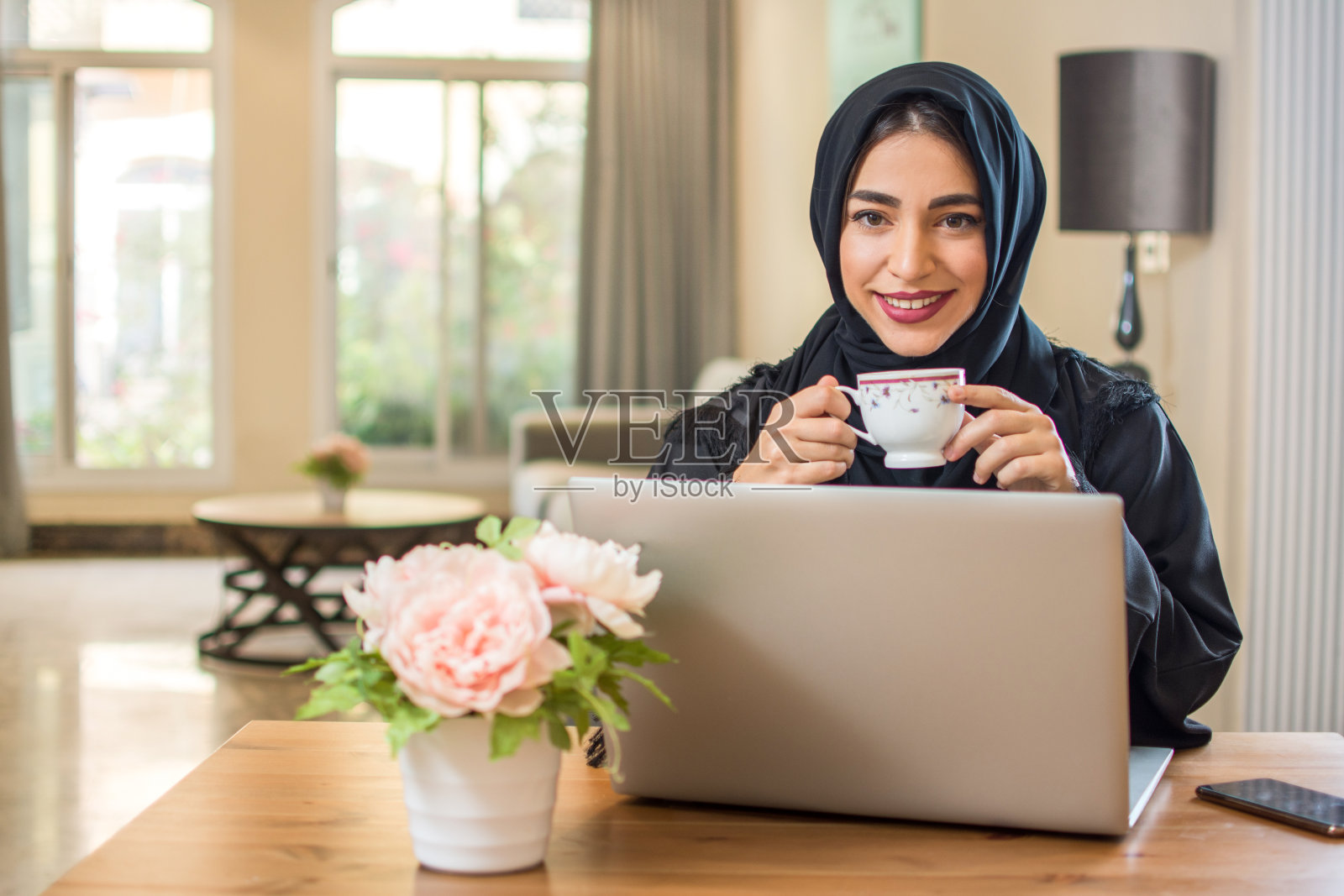 年轻的阿拉伯妇女拿着咖啡杯，用笔记本电脑看着摄像头照片摄影图片