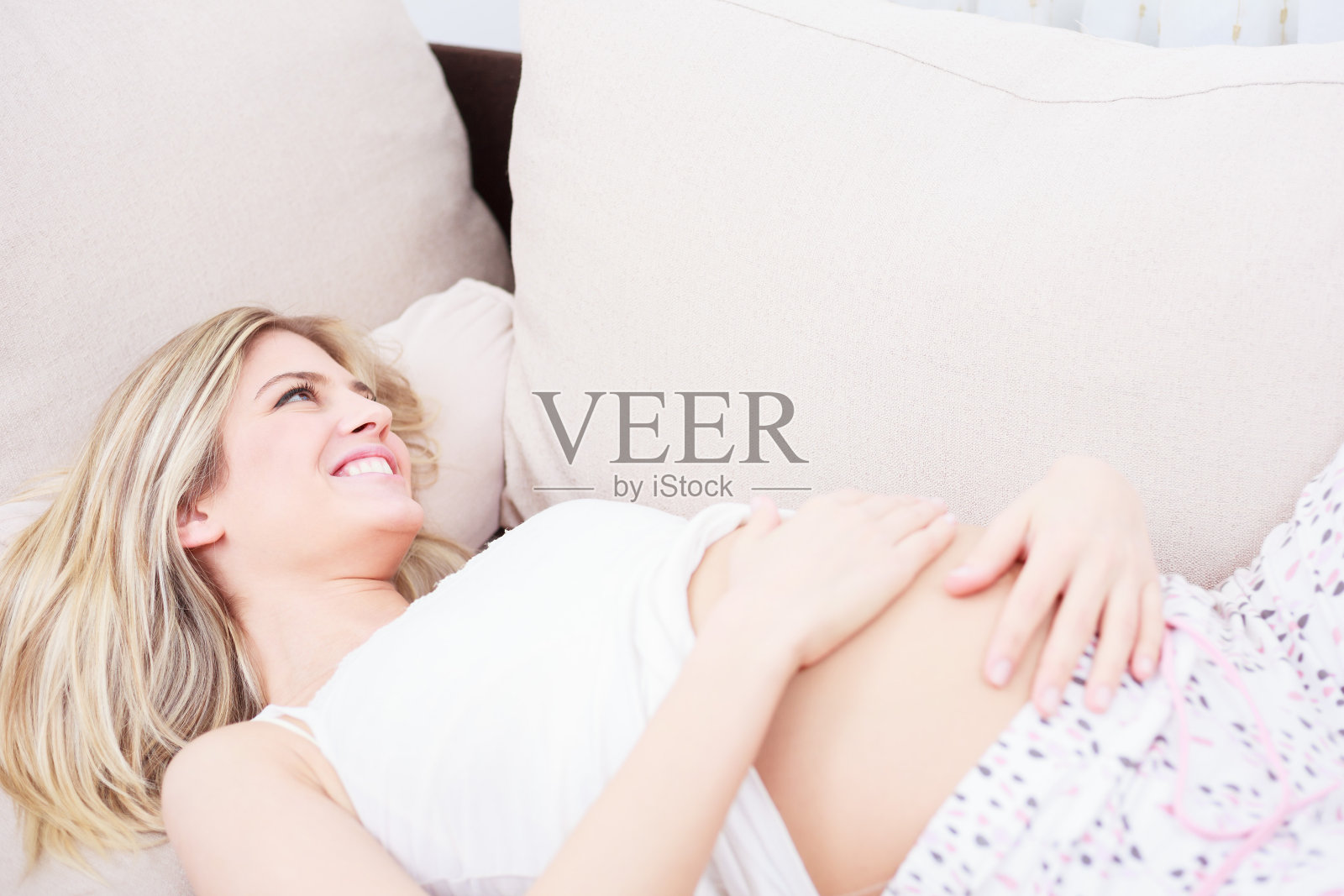 快乐的怀孕年轻女子微笑着，手臂放在她的腹部，而躺在沙发上照片摄影图片