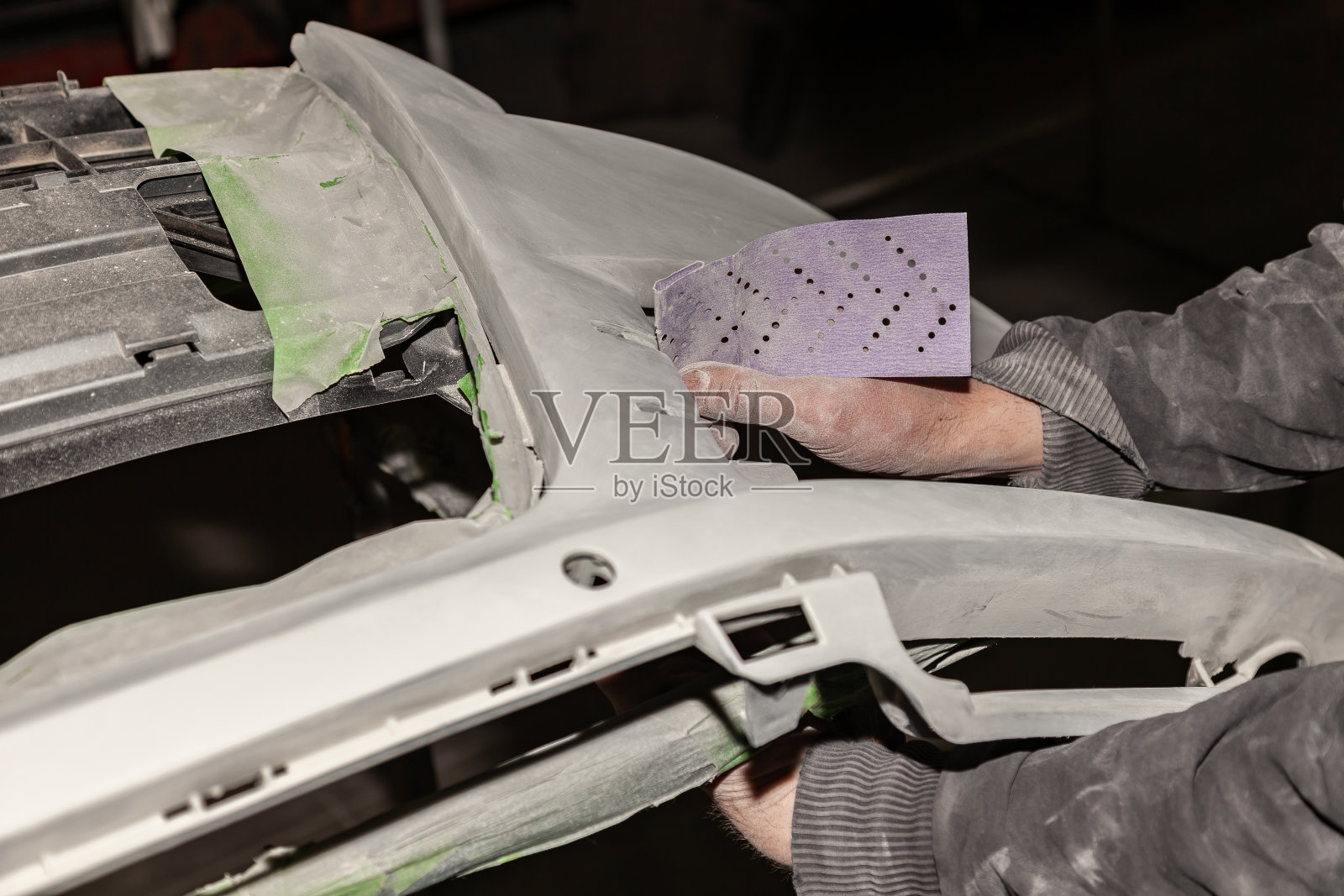 在汽车修理厂，一名男子在打磨纸的帮助下，准备了一辆车的车身部件，以备涂装之用。碰撞后的恢复保险杠。照片摄影图片