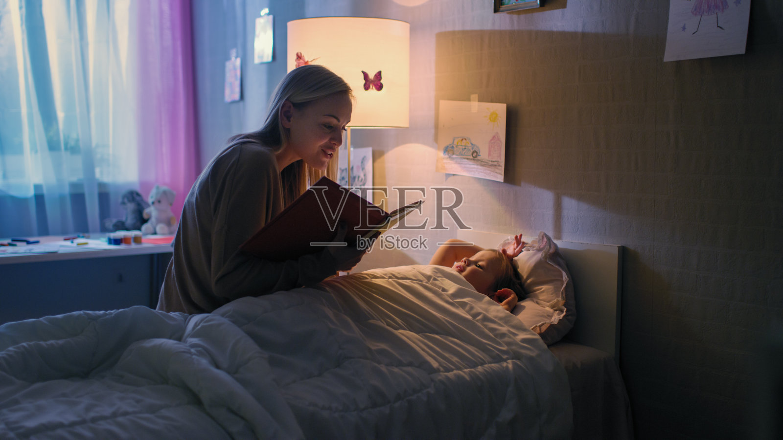 年轻慈爱的母亲读睡前故事给她美丽的小女儿，她在她的床上睡觉。照片摄影图片