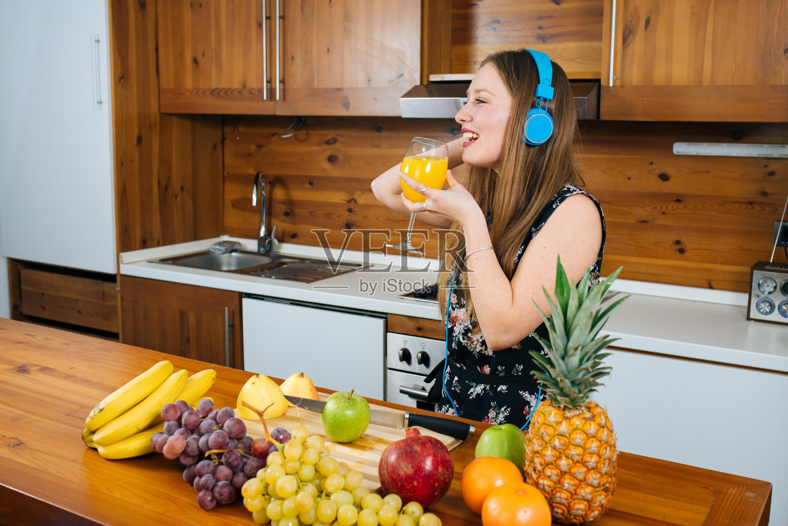 漂亮的女孩戴着耳机，拿着电话在厨房里喝着橙汁。照片摄影图片