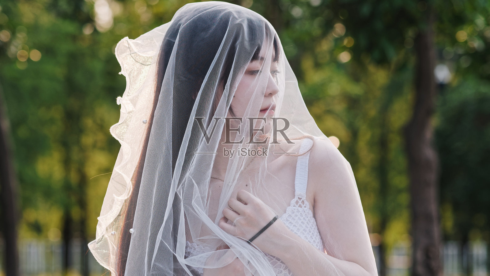 美丽的中国女孩的肖像在白色的新娘礼服看着远离与头面纱在风黄昏模糊的森林背景，美丽，生活方式和夏天的概念。照片摄影图片