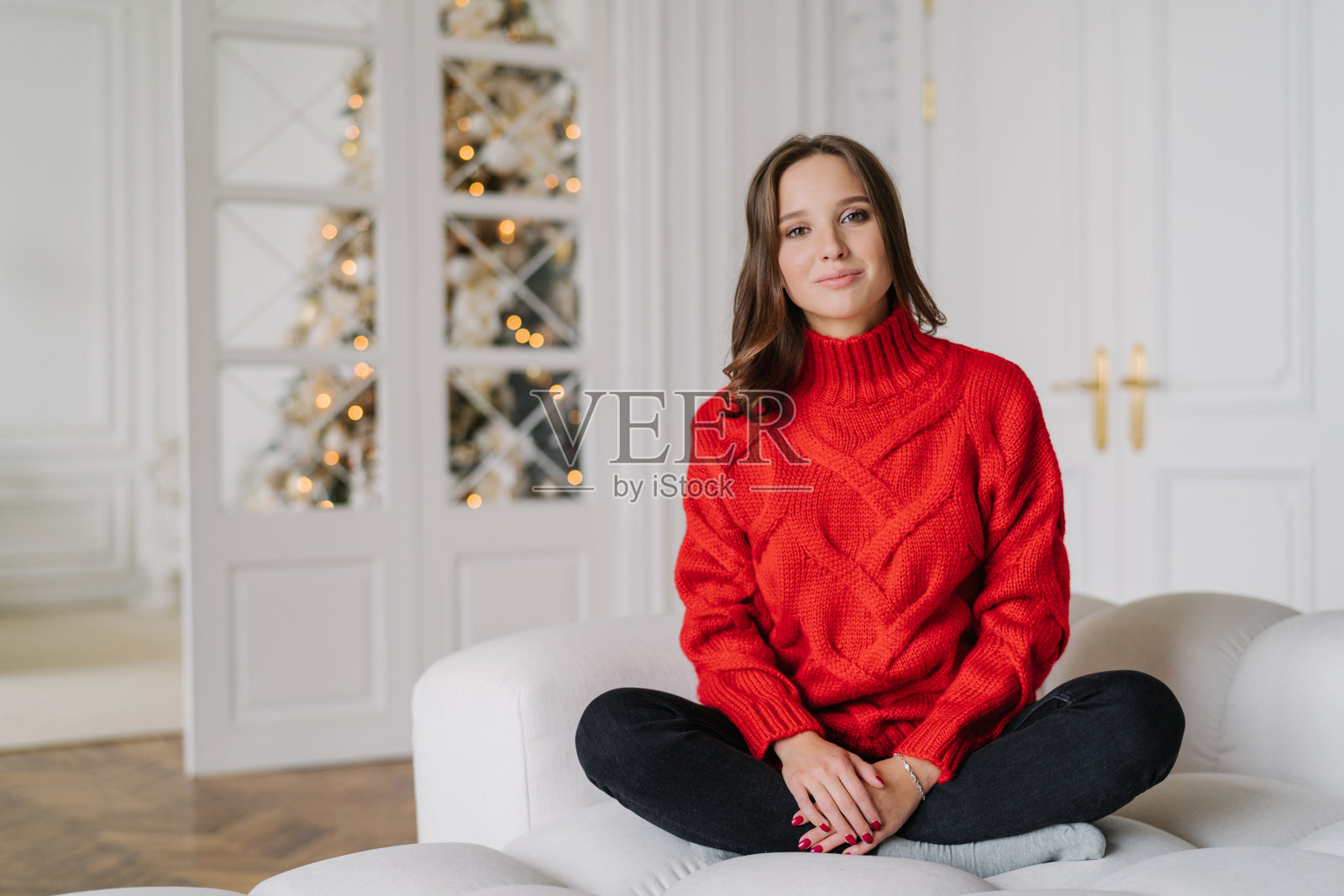 照片上的年轻女子穿着红色的毛衣、裤子，交叉着双腿坐在舒适的沙发上，享受着家庭气氛，背景是装饰好的新年树。家的概念照片摄影图片