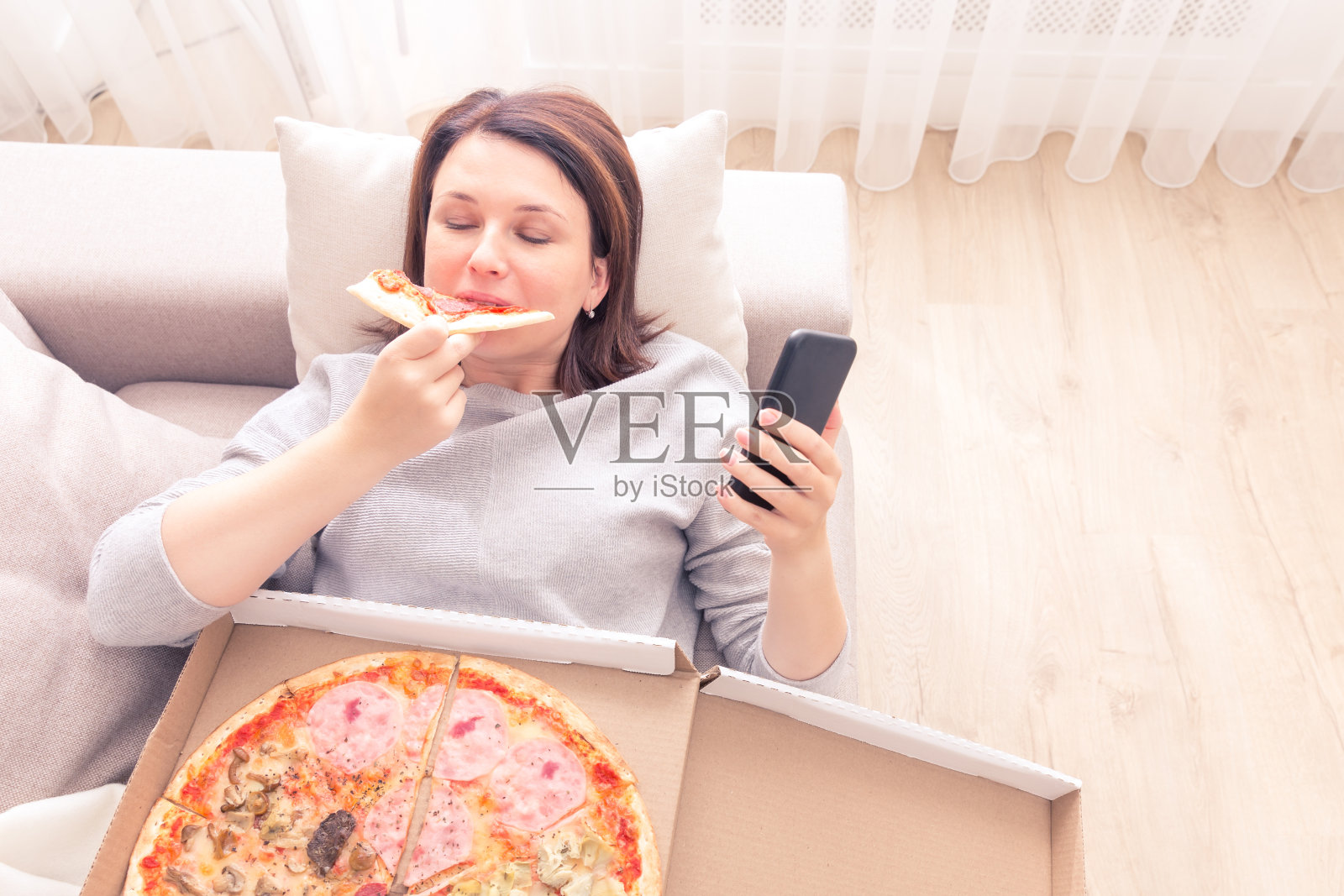 伤心的女人躺在家里的沙发上吃着披萨拿着电话，暖暖的黄色调照片摄影图片