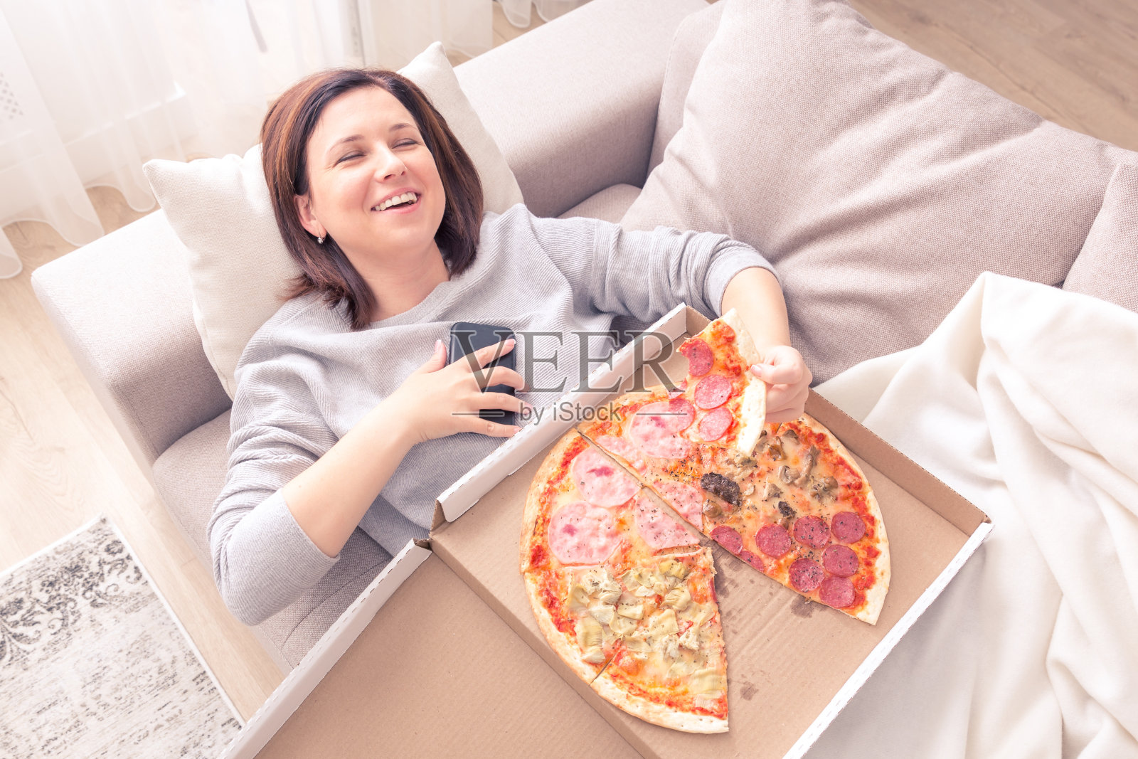 一个笑着的年轻女人的肖像躺在沙发上，吃着披萨，拿着电话橙色音照片摄影图片