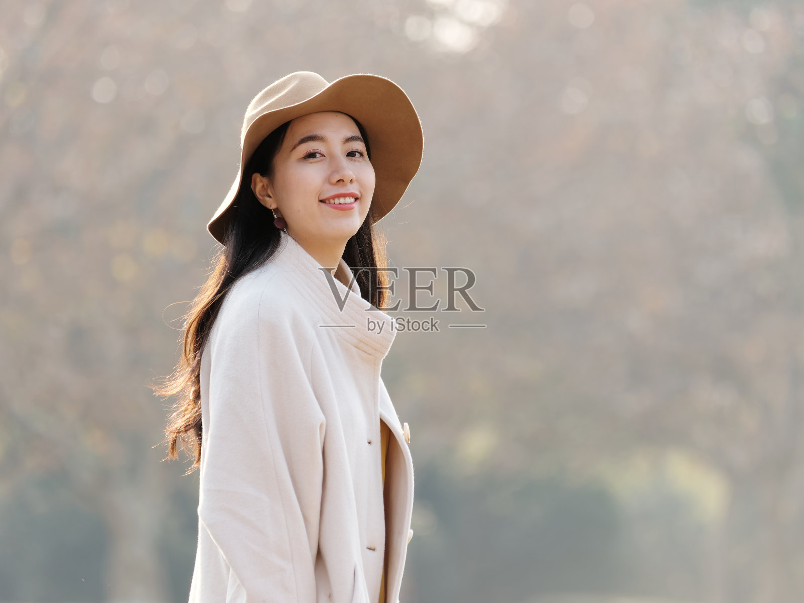 美丽的中国年轻女子在米色外套和帽子摆姿势和微笑的镜头，迷人的中国女孩与黑色的长发享受她的休闲户外时间。照片摄影图片