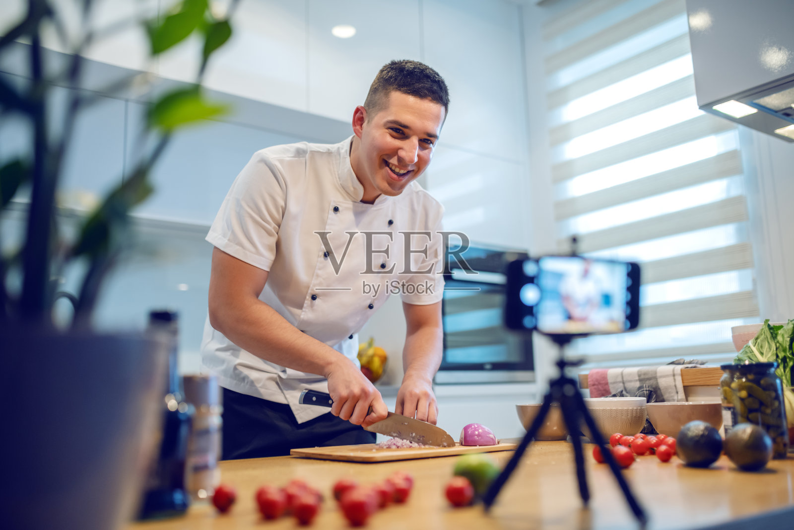 微笑的白人厨师穿着制服站在厨房切洋葱，同时记录自己的博客。厨房柜台上放着蔬菜和香料。照片摄影图片