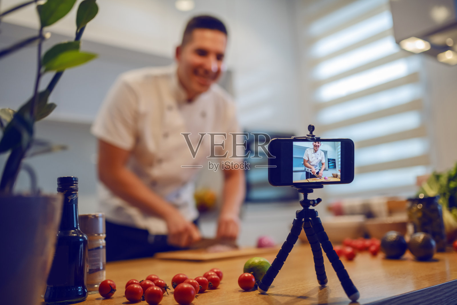 微笑的白人厨师穿着制服站在厨房切洋葱，同时拍摄自己的博客。厨房柜台上放着蔬菜和香料。选择性地专注于智能手机。照片摄影图片