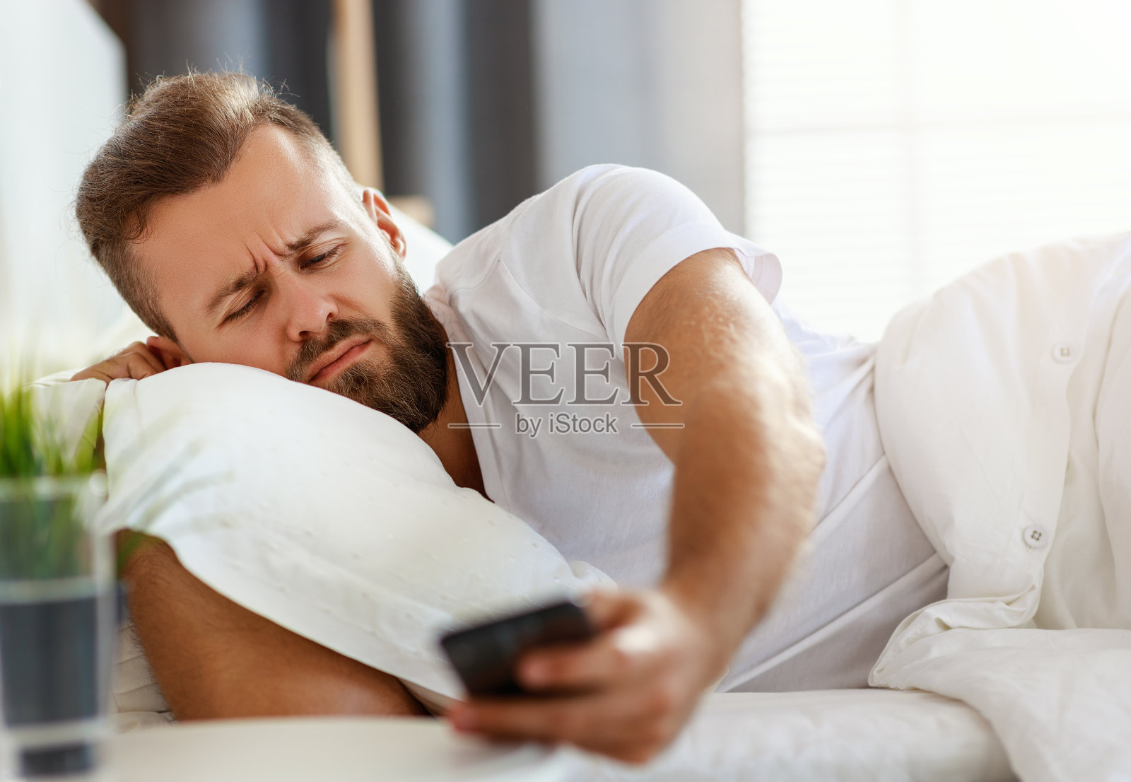 年轻的大胡子男人早上在他的床上用手机闹钟醒来照片摄影图片