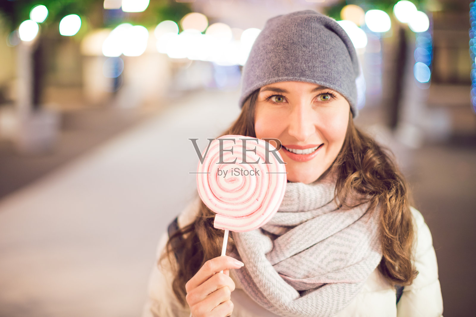 在意大利的圣诞街道上，一个年轻快乐的女人拿着一个棉花糖棒棒糖(棒棒糖)在她的手作为背景照片摄影图片