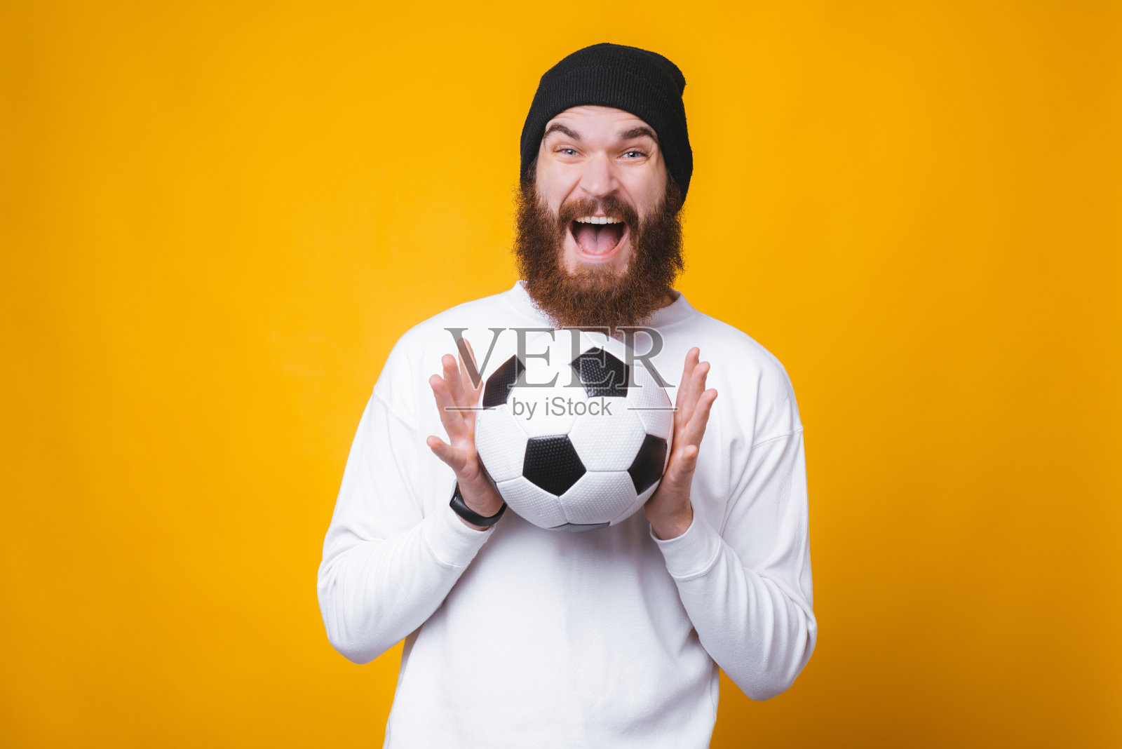 黄色的墙上，快乐的大胡子兴奋地拿着一个足球。照片摄影图片
