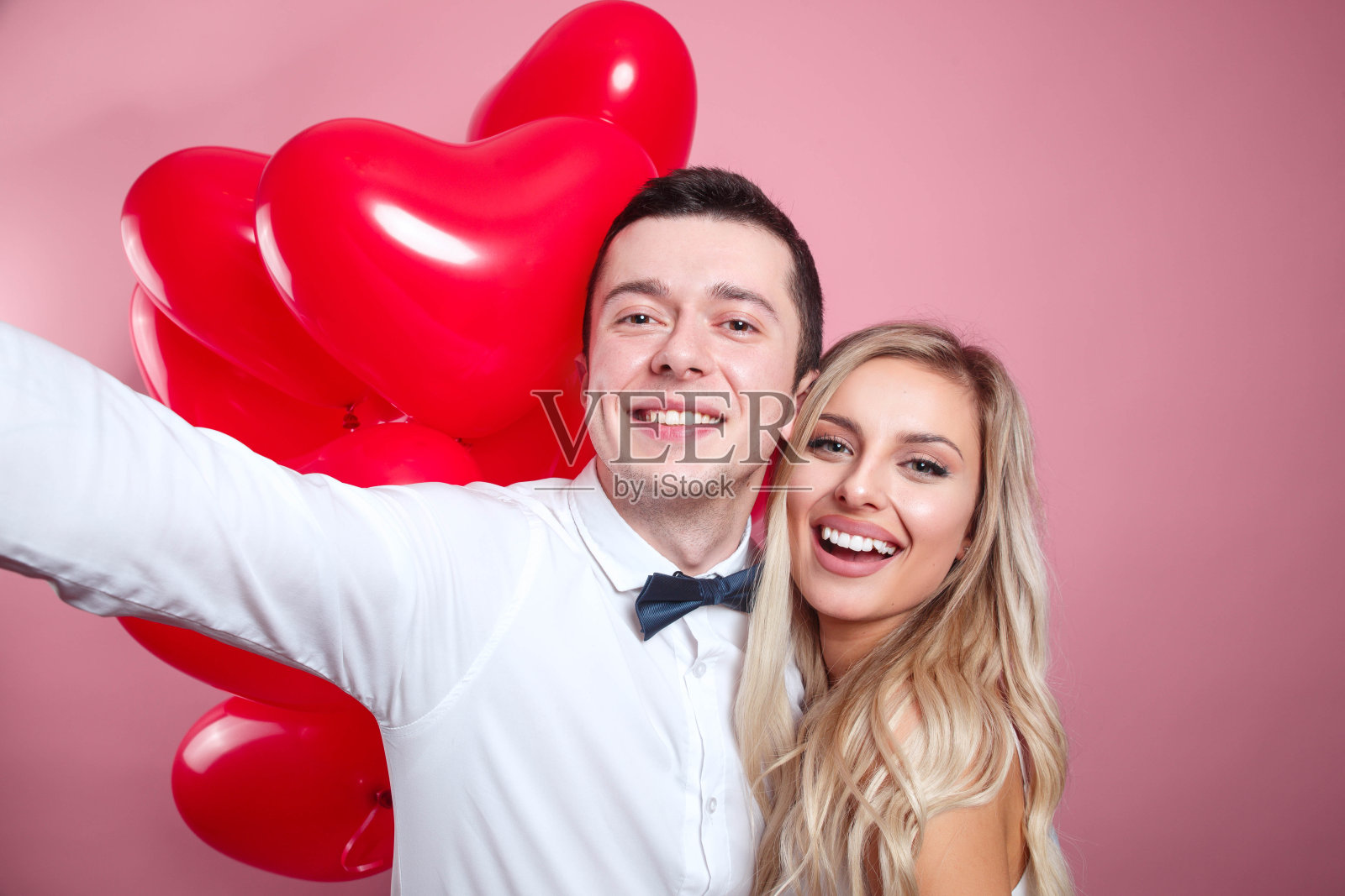 一对美丽的年轻夫妇用手机自拍，粉色背景上站着红色气球照片摄影图片