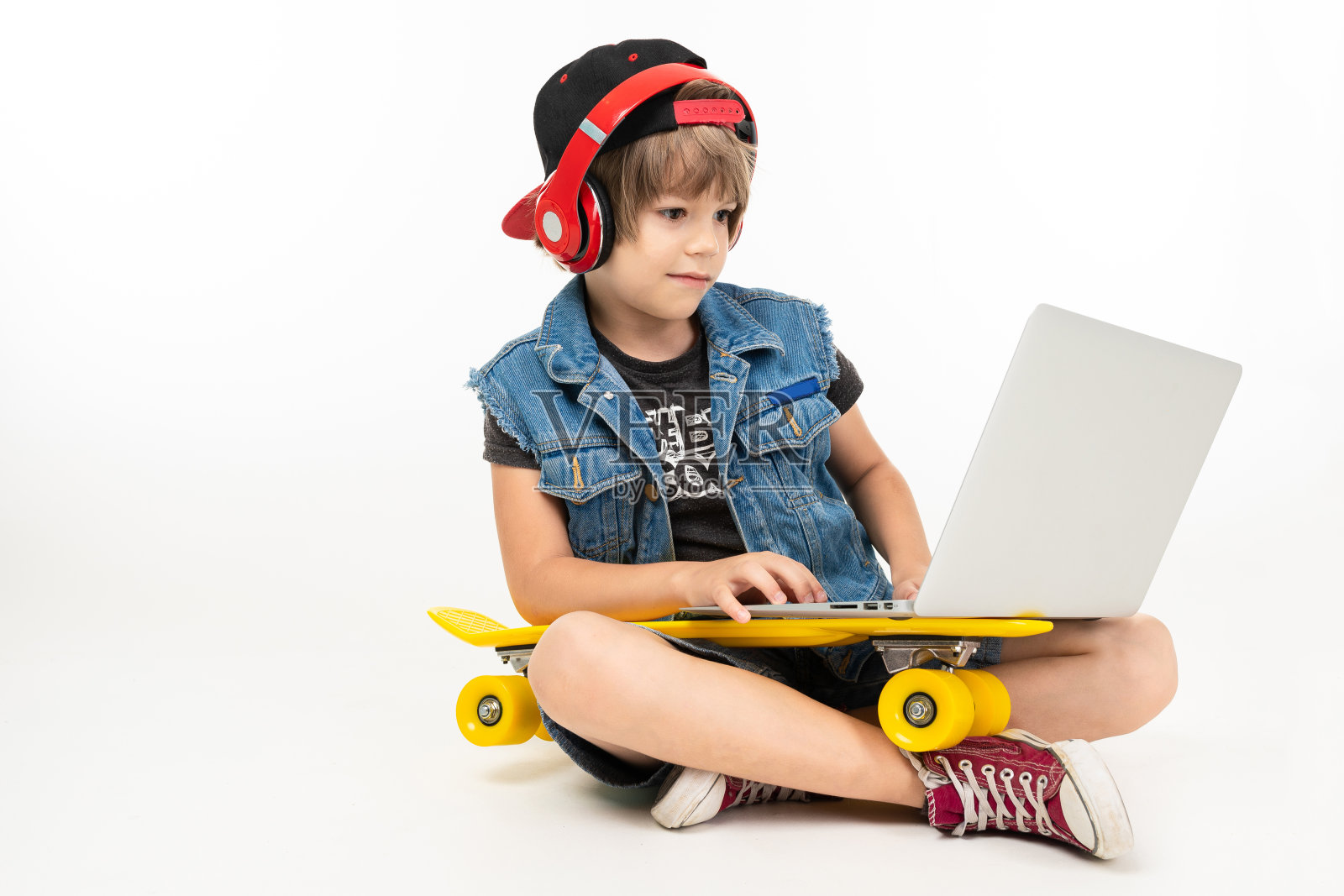 十几岁的男孩坐在地板上，穿着牛仔夹克和短裤。黄色便士运动鞋，红色耳机和笔记本电脑隔离在白色背景照片摄影图片