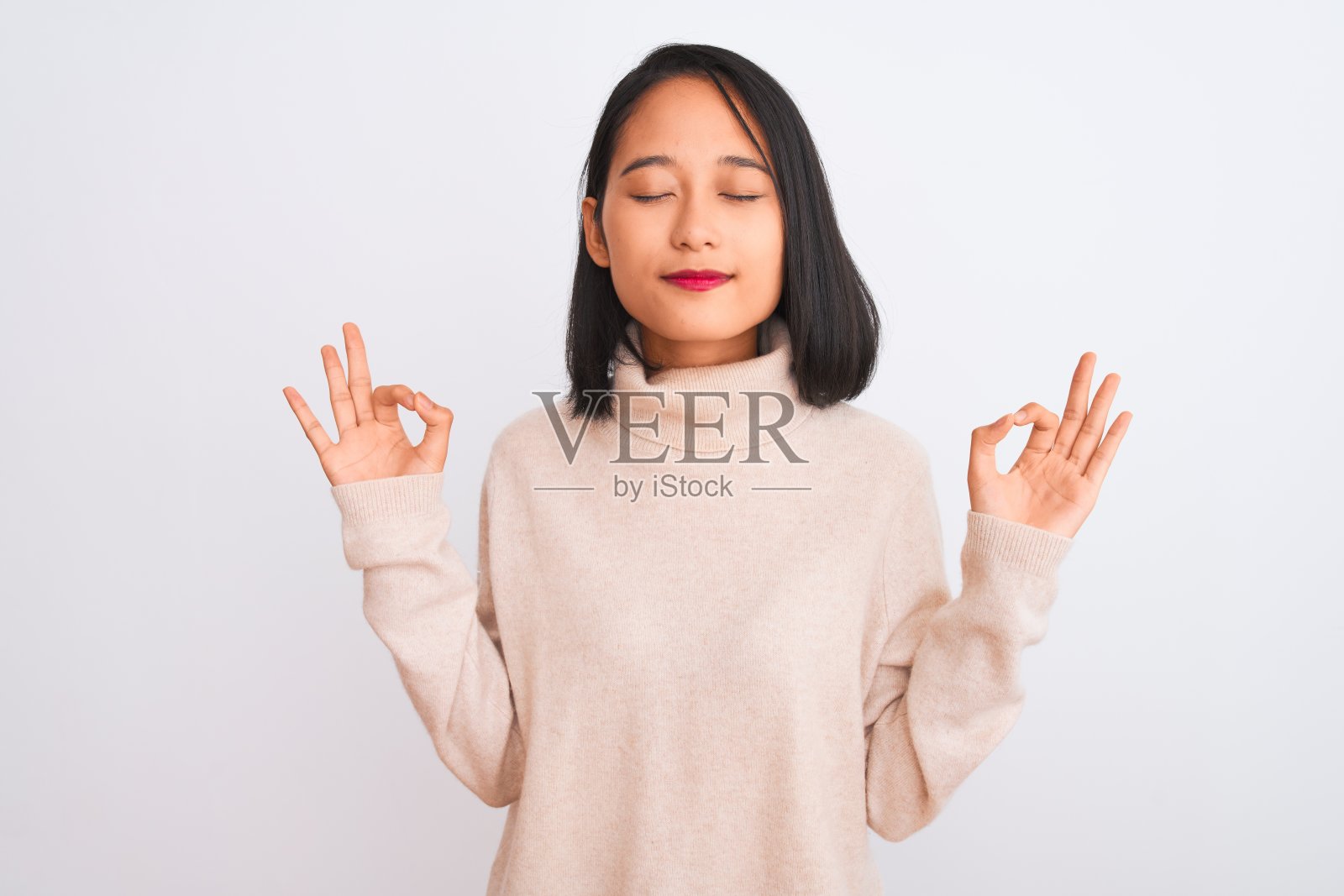 年轻的中国妇女穿着高领毛衣站在孤立的白色背景放松和微笑，闭着眼睛做冥想手势与手指。瑜伽的概念。照片摄影图片