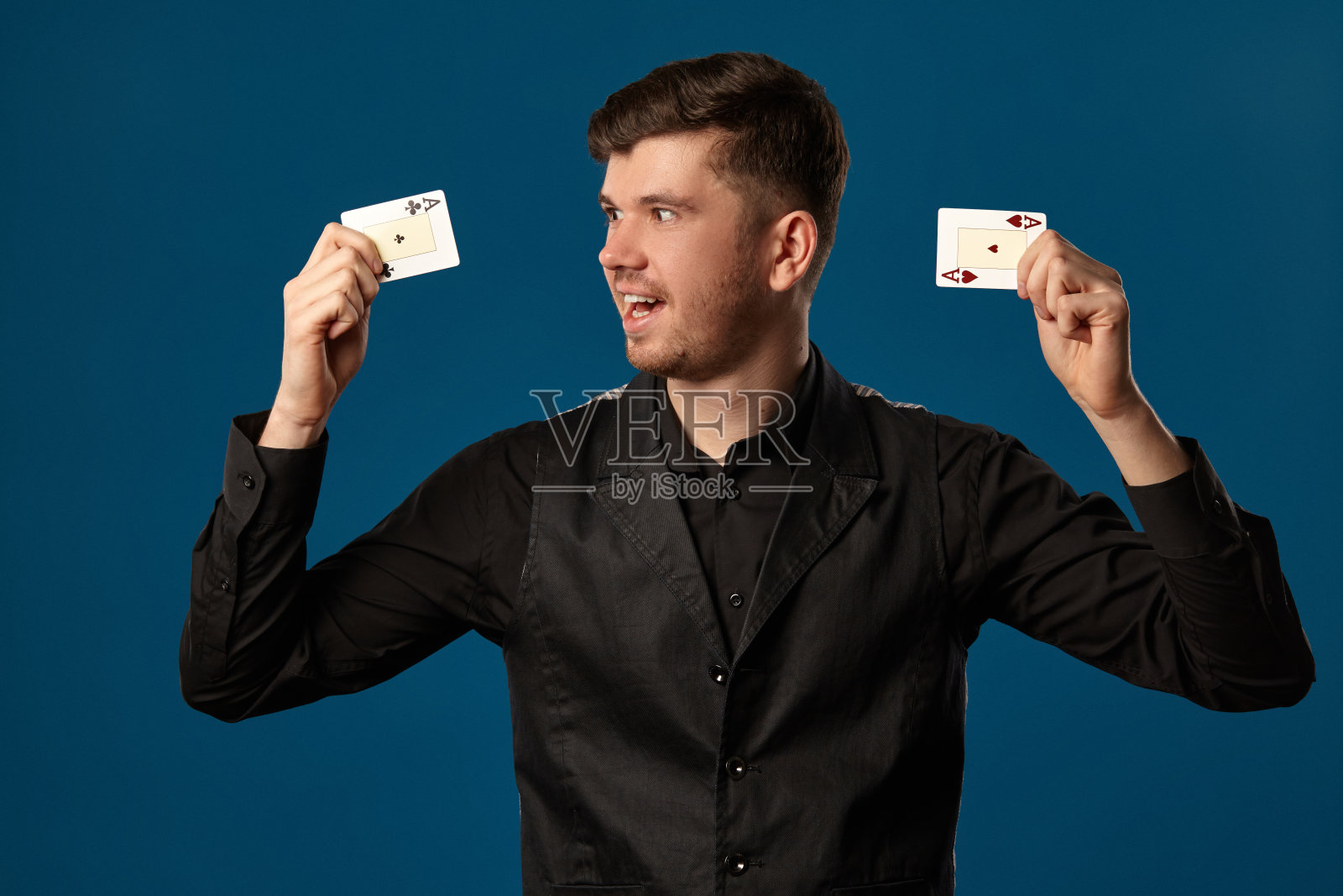 玩扑克的新手，穿着黑色背心和衬衫。拿着两张扑克牌，在蓝色的工作室背景下摆姿势。赌博,赌场。特写镜头照片摄影图片