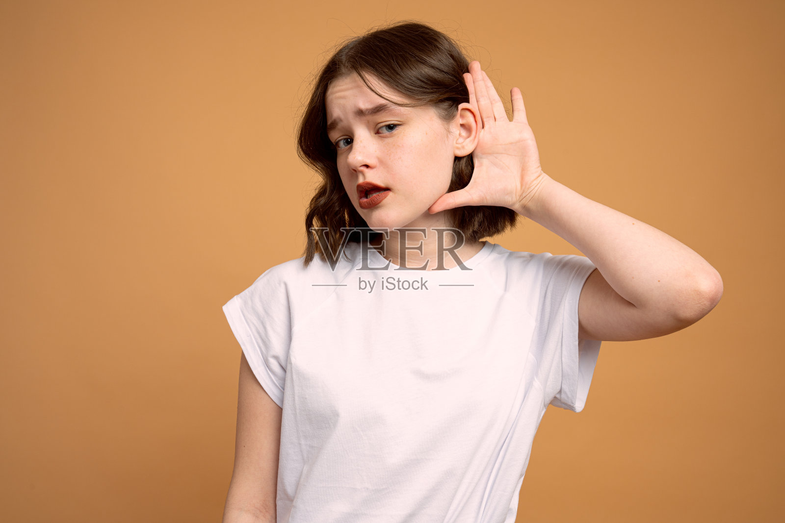 年轻的黑发女孩在白色t恤八卦与手靠近她的耳朵隔离在橙色背景照片摄影图片